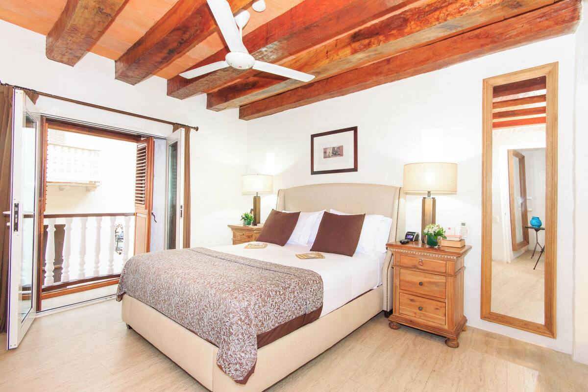 Une chambre à coucher comprenant un lit, des lampes de chevet, une commode, un ventilateur de plafond, un tableau encadré et une porte-fenêtre coulissante donnant sur un balcon.