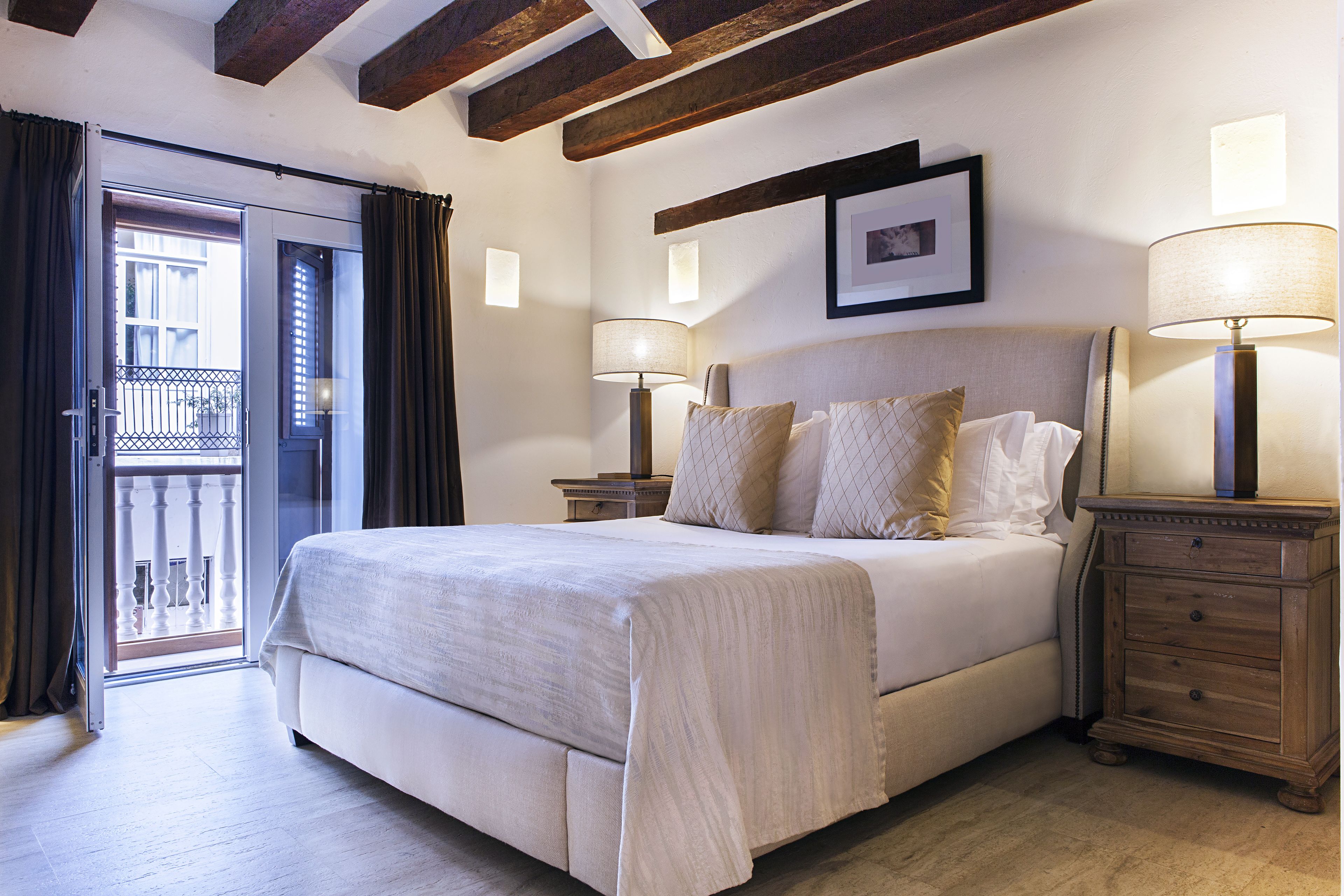 A bedroom with a large bed, two bedside tables with lamps, a wooden nightstand, a framed picture on the wall, and a balcony door.