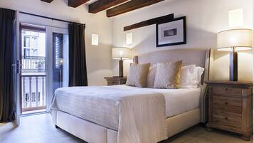 A bedroom with a large bed, two bedside tables with lamps, a wooden nightstand, a framed picture on the wall, and a balcony door.