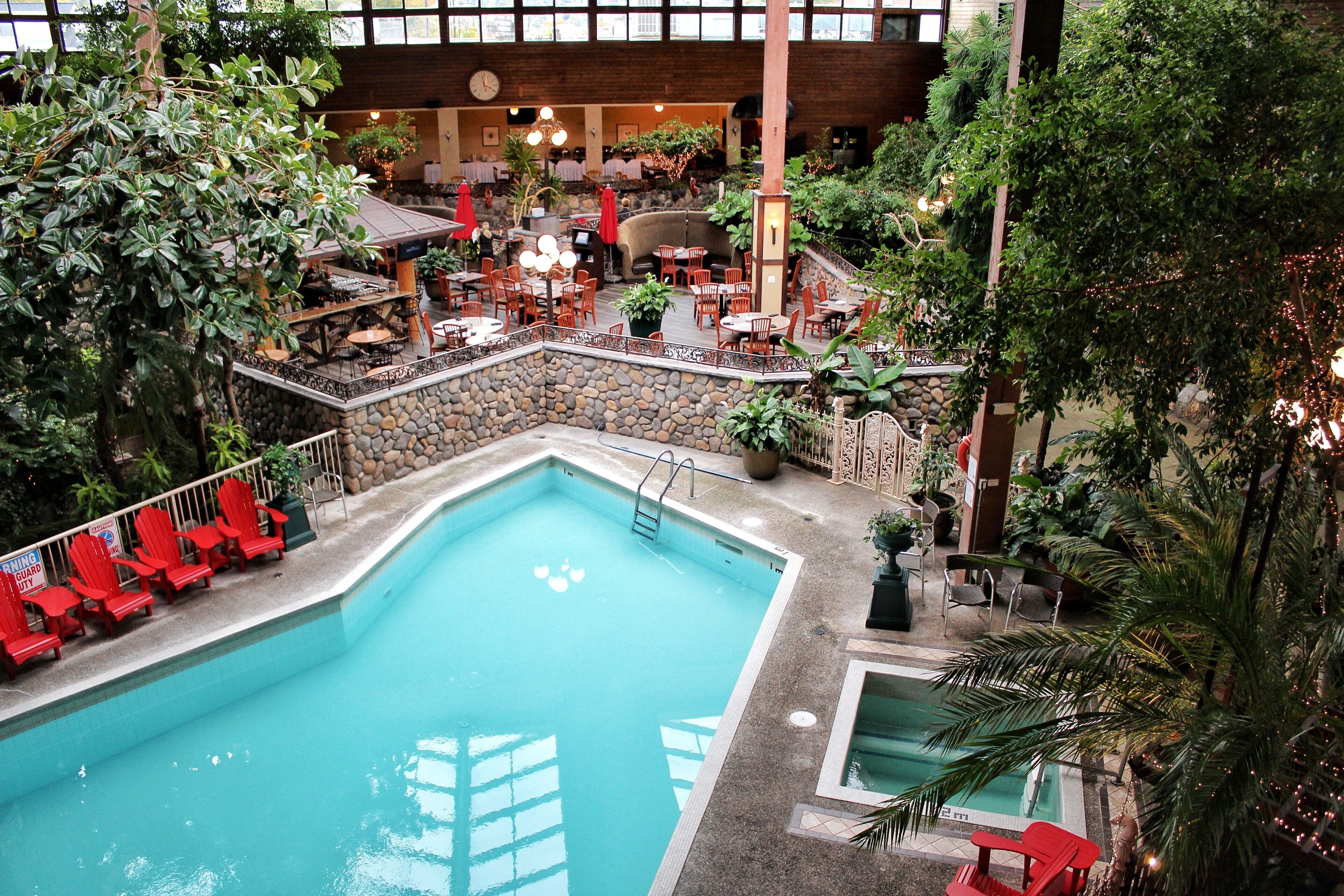An indoor pool with red chairs and tables, surrounded by greenery and stone walls.