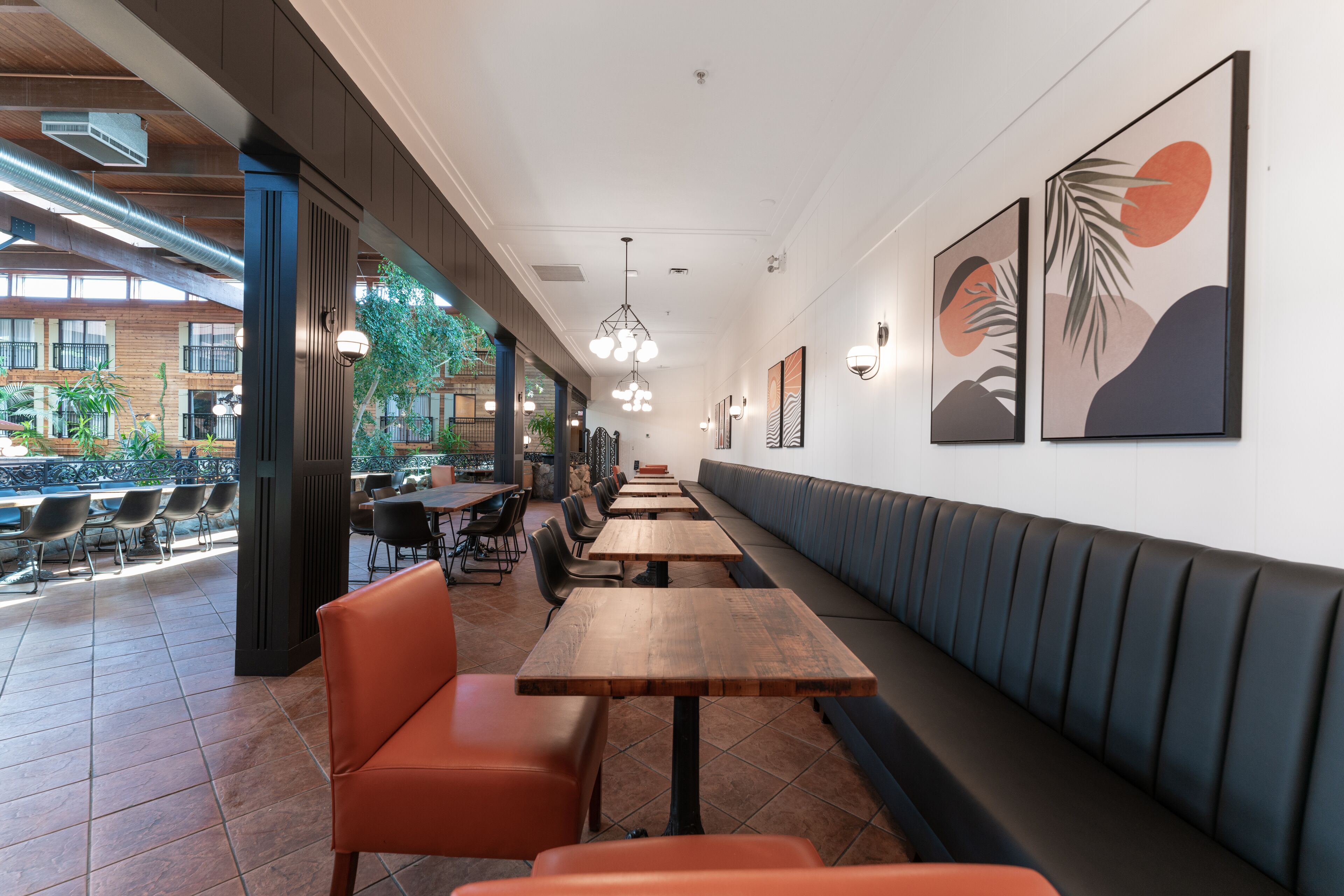 Een restaurant met lange houten tafels en oranje stoelen, een zwarte bank met kussens langs de muur, en lijstwerken aan de wanden.