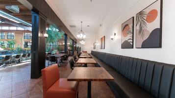 Een restaurant met lange houten tafels en oranje stoelen, een zwarte bank met kussens langs de muur, en lijstwerken aan de wanden.