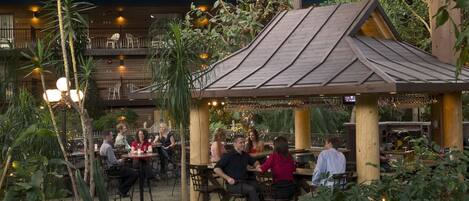An outdoor dining area with tables and chairs, surrounded by plants and a wooden structure.