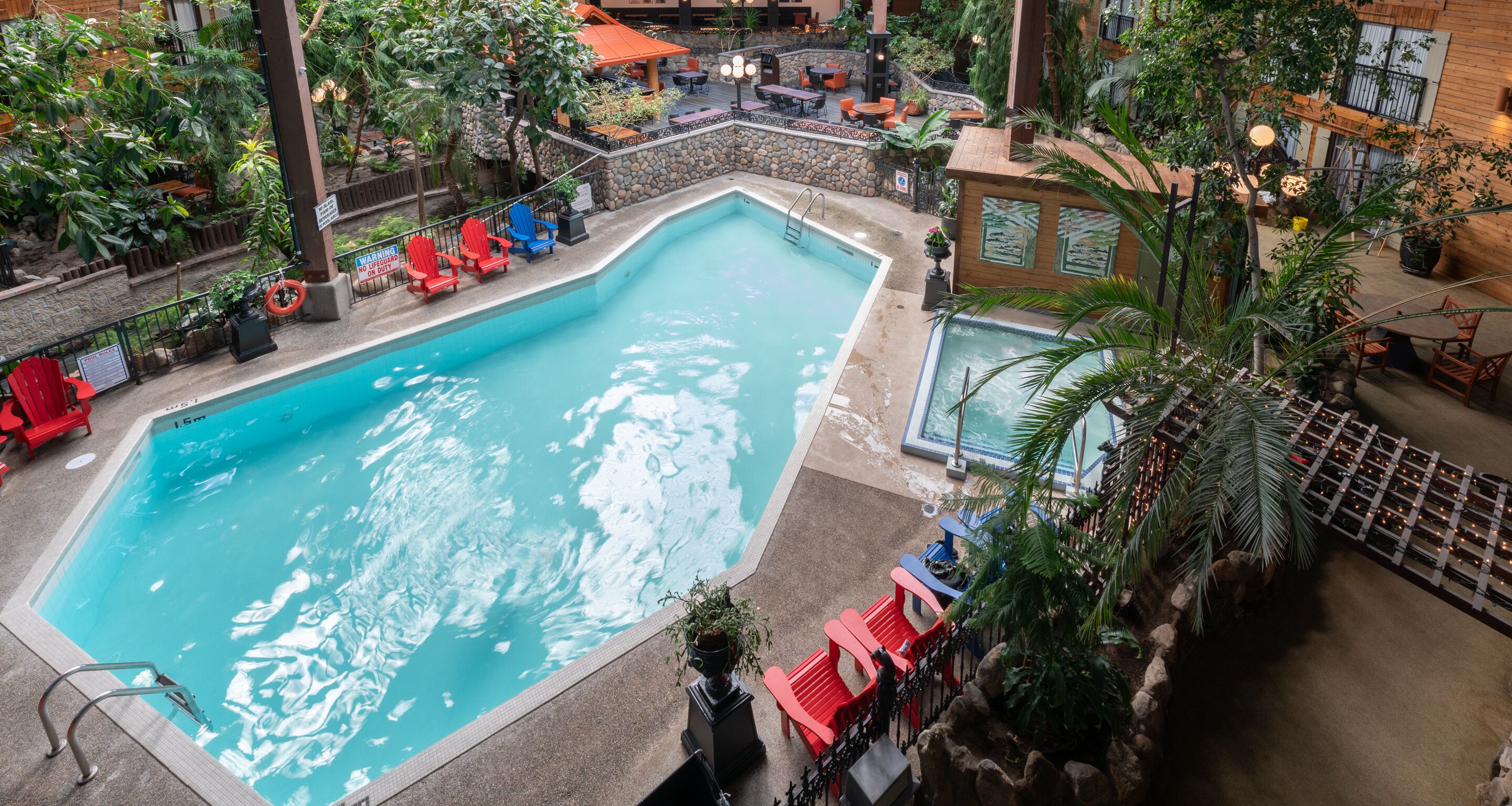 An indoor swimming pool with a stone border, surrounded by red and blue chairs, and various plants.
