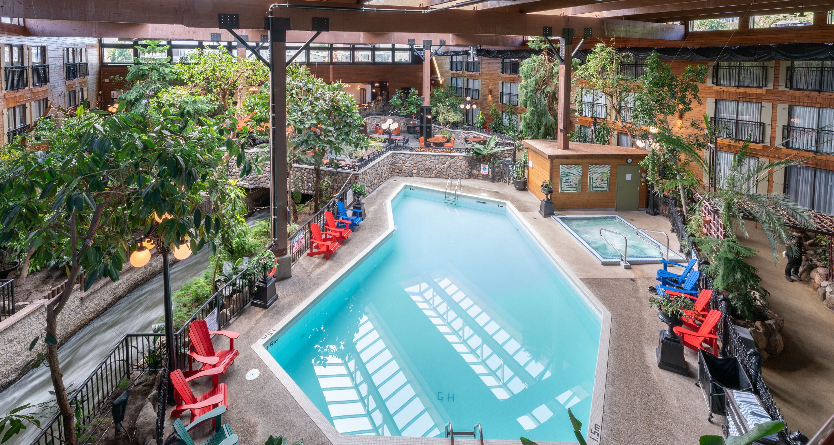An indoor swimming pool with red and blue lounge chairs, surrounded by greenery and stone walls.