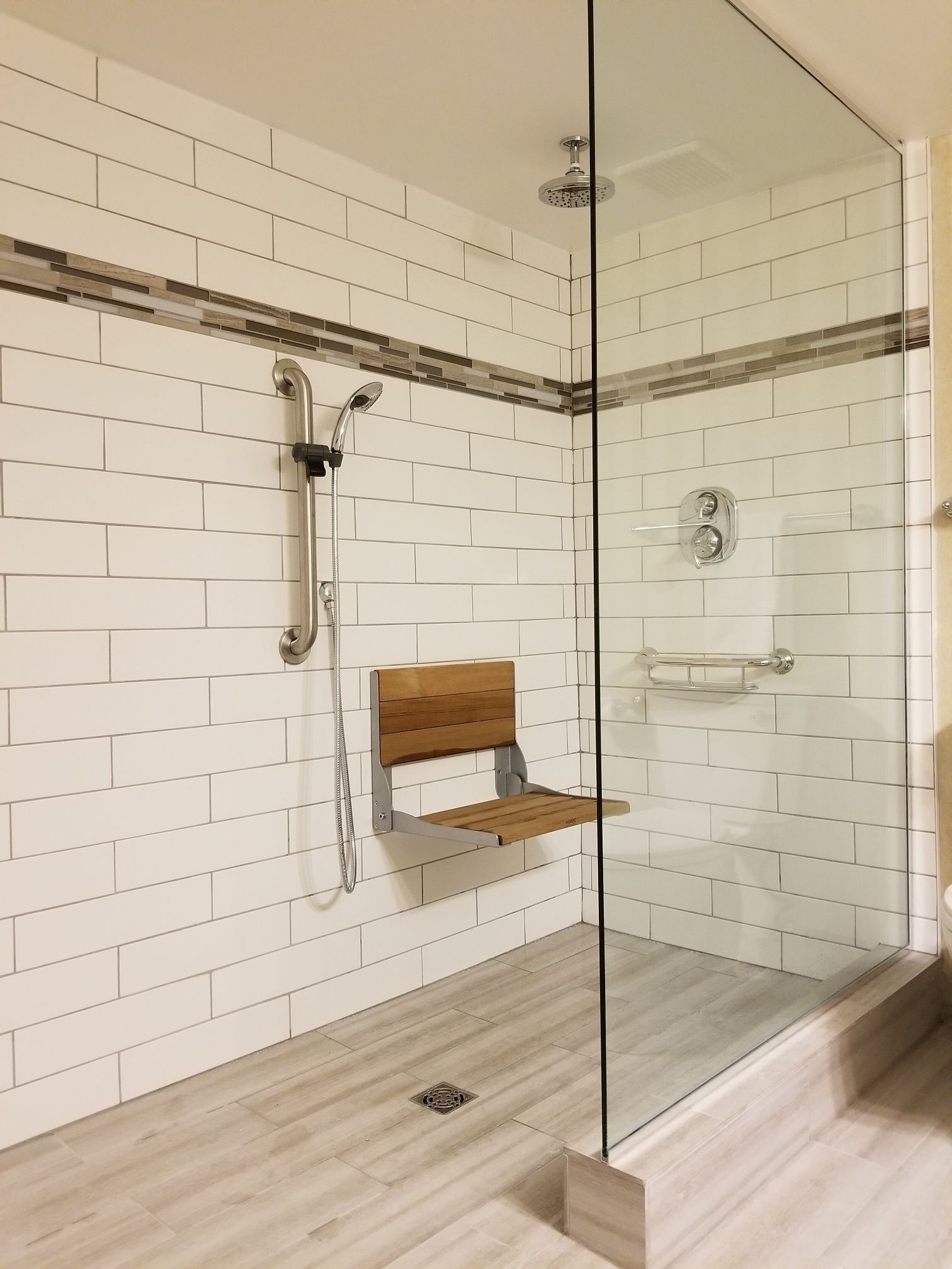 A modern shower with a glass door, white tiled walls, and a wooden bench.