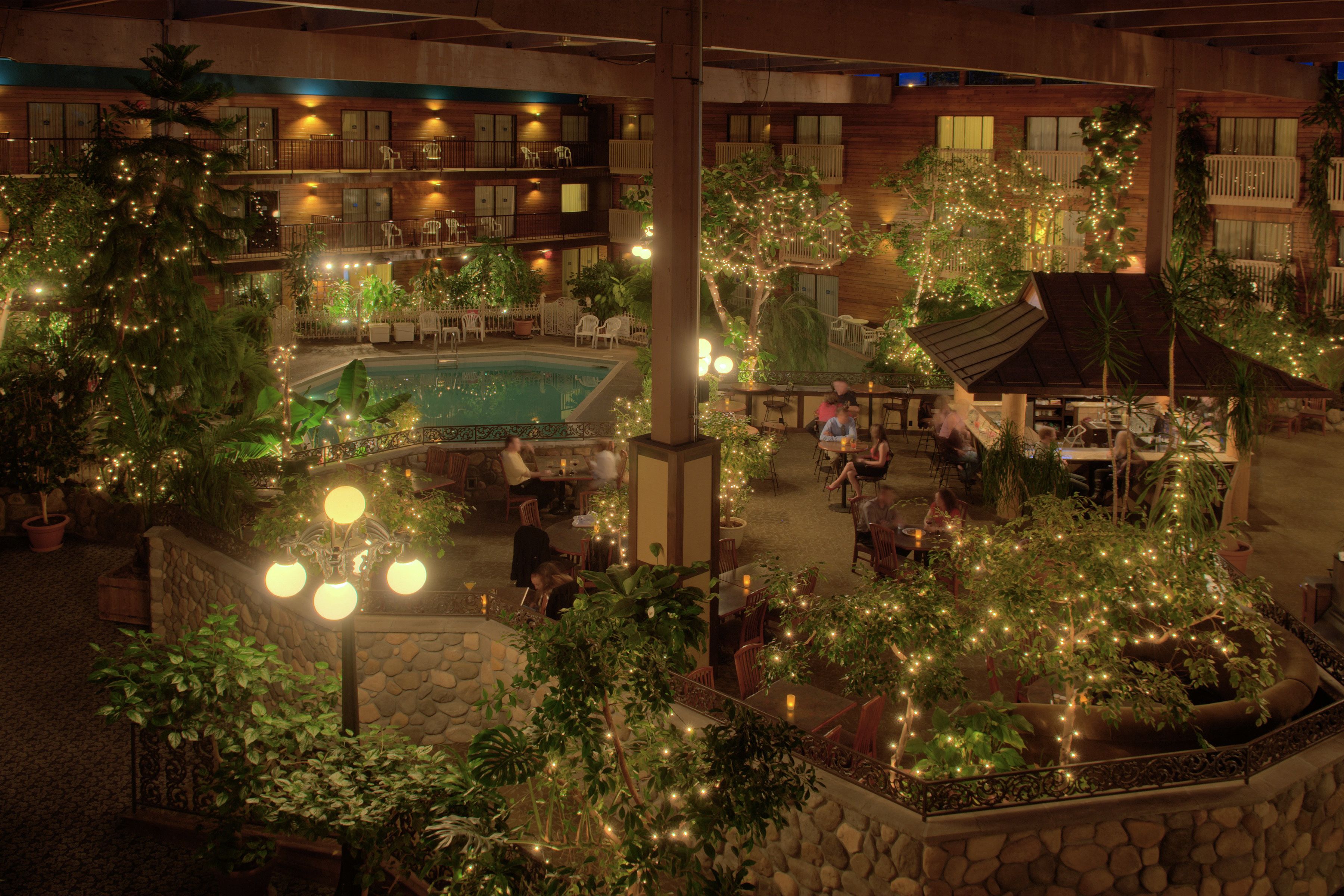An indoor courtyard with a pool, surrounded by potted plants and trees, and multiple dining areas with tables and chairs.