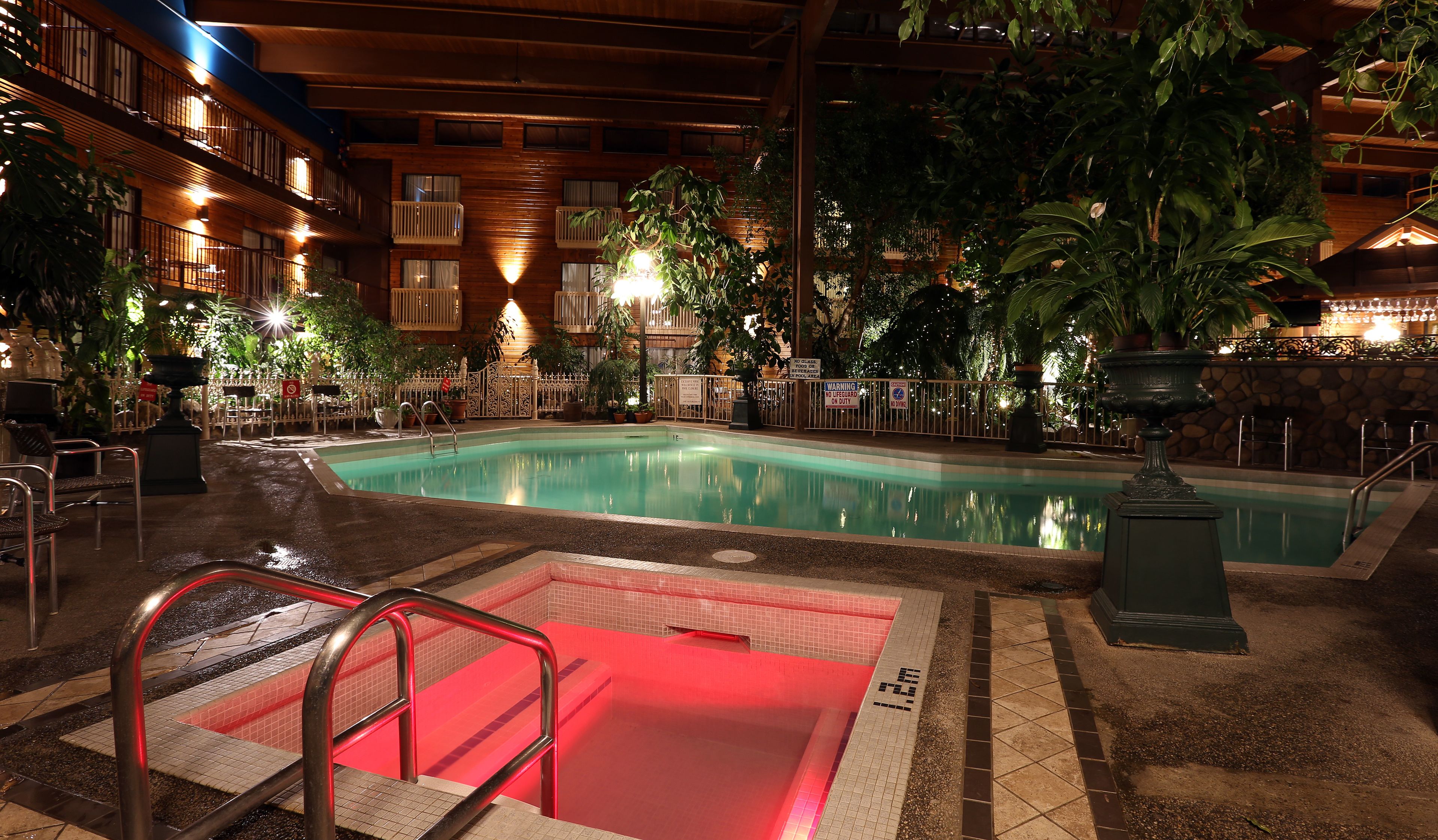 An indoor pool area with a red-lit jacuzzi, surrounded by potted plants and a wooden deck.