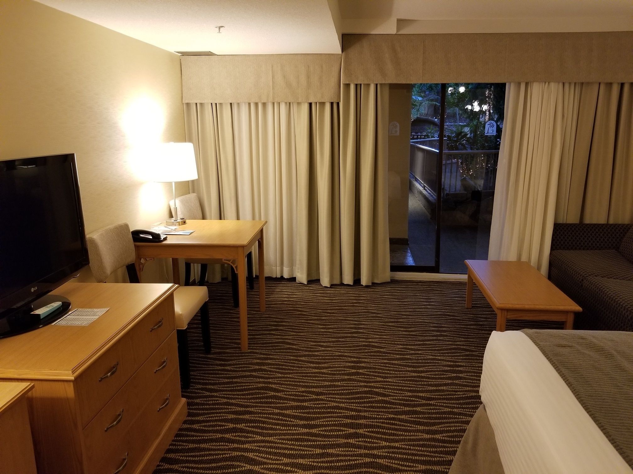 Ein Hotelzimmer mit Bett, Schreibtisch mit Lampe, Fernseher und Blick ins Freie durch eine Schiebetür.
