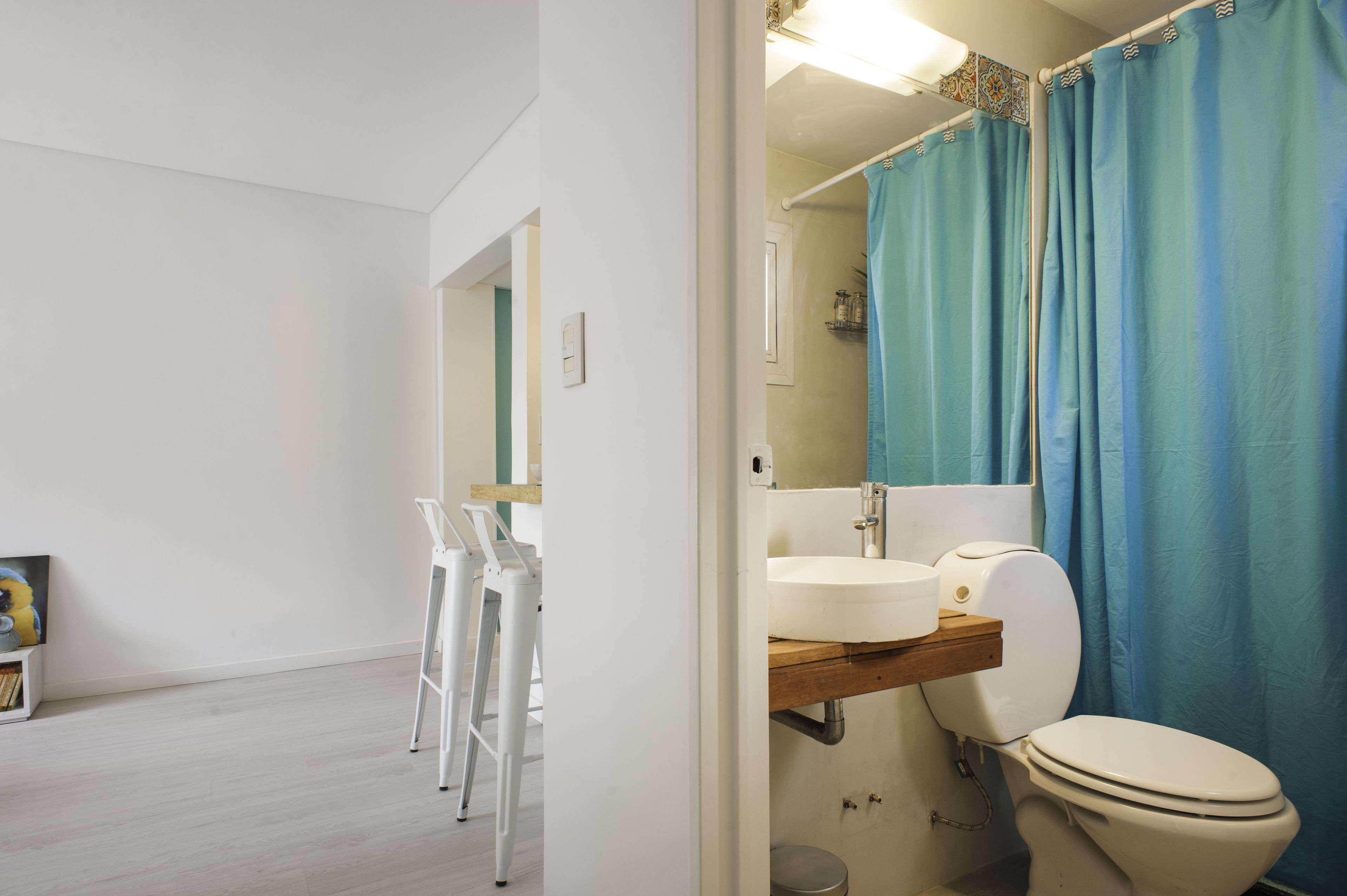 A bathroom with a white sink, a toilet, and a blue shower curtain. There is a wooden shelf above the sink and a white ladder leaning against the wall.
