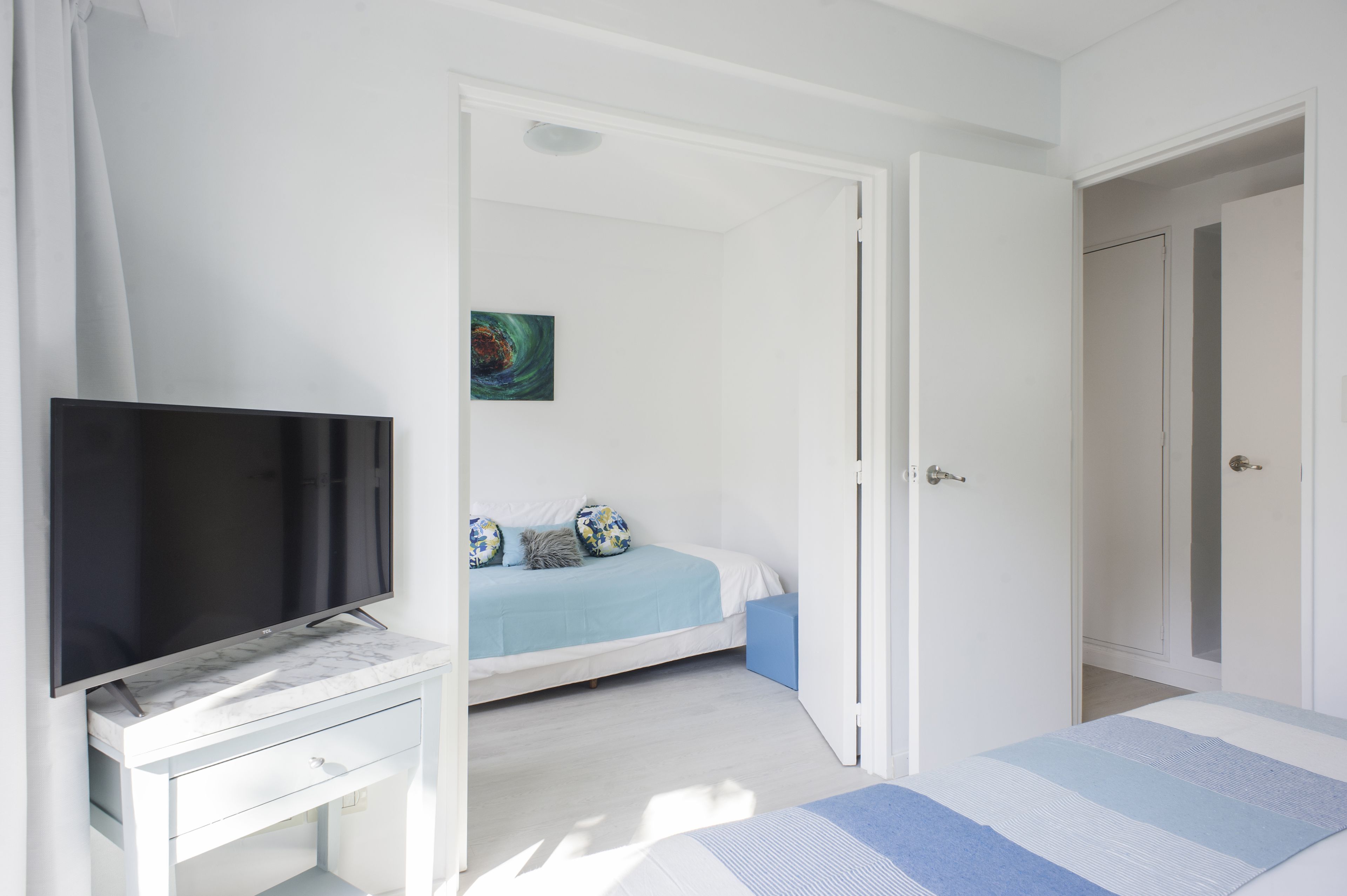 A bedroom with a bed, a TV on a desk, and a door leading to another room.