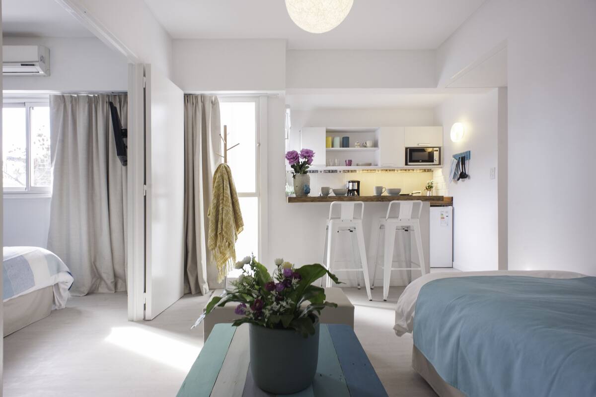 A modern bedroom with a bed, a bedside table with a potted plant, and a view into a kitchen area with a dining table and chairs.