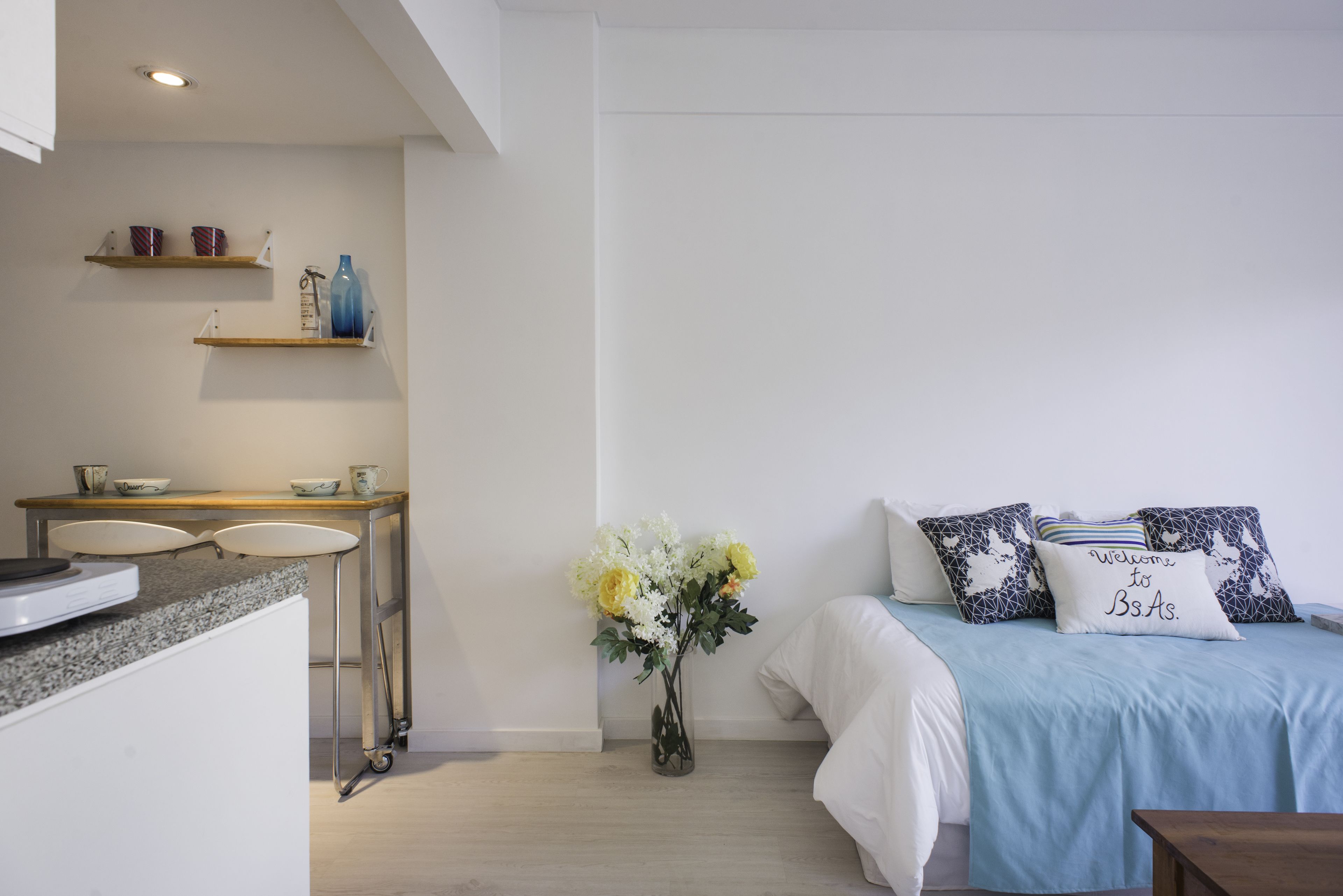 A modern bedroom with a bed, bedside table, and a small kitchenette area with a sink and shelves.