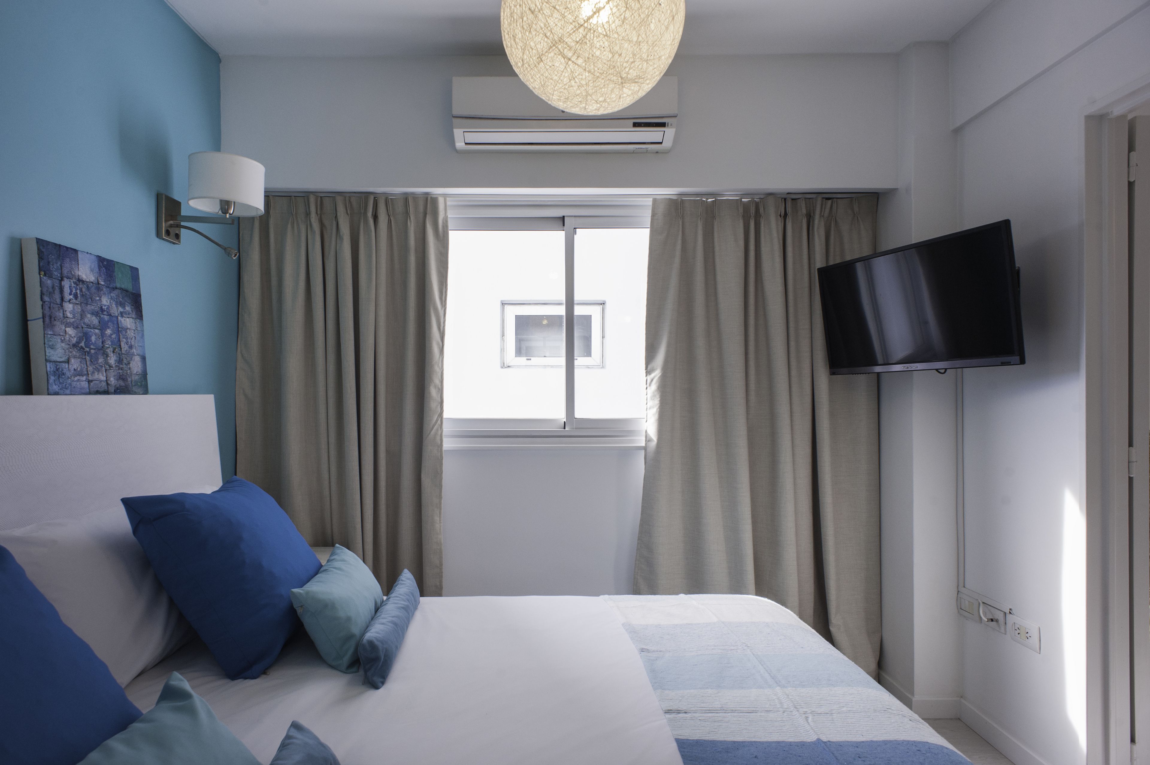 A bedroom with a bed, a flat-screen TV, an air conditioning unit, a window with curtains, and a wall-mounted lamp.