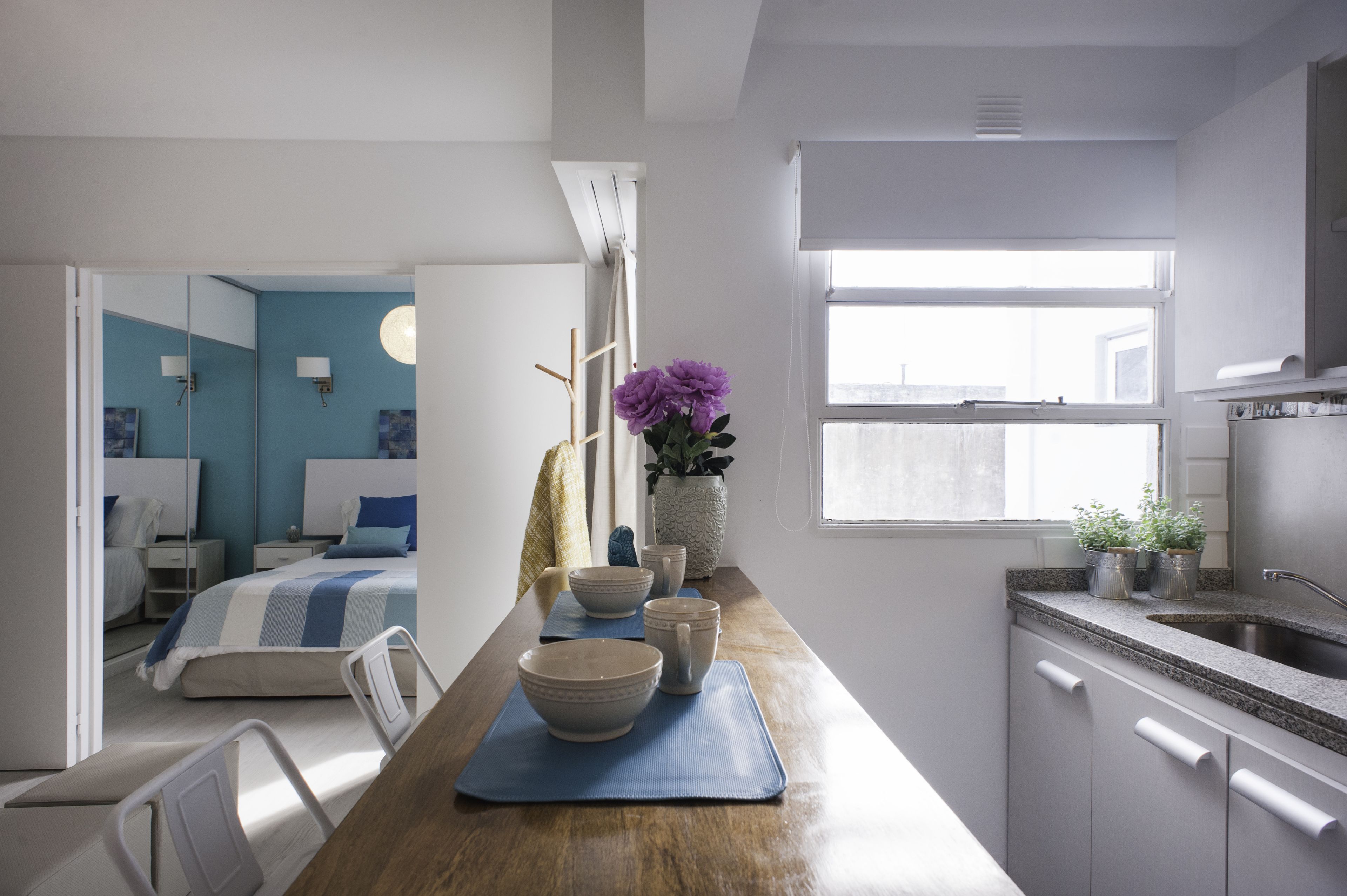 Habitación de hotel moderna con una cama grande, mesitas de noche, una pequeña zona de estar, una kitchenette y una ventana que deja entrar luz natural.