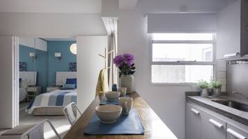 Habitación de hotel moderna con una cama grande, mesitas de noche, una pequeña zona de estar, una kitchenette y una ventana que deja entrar luz natural.