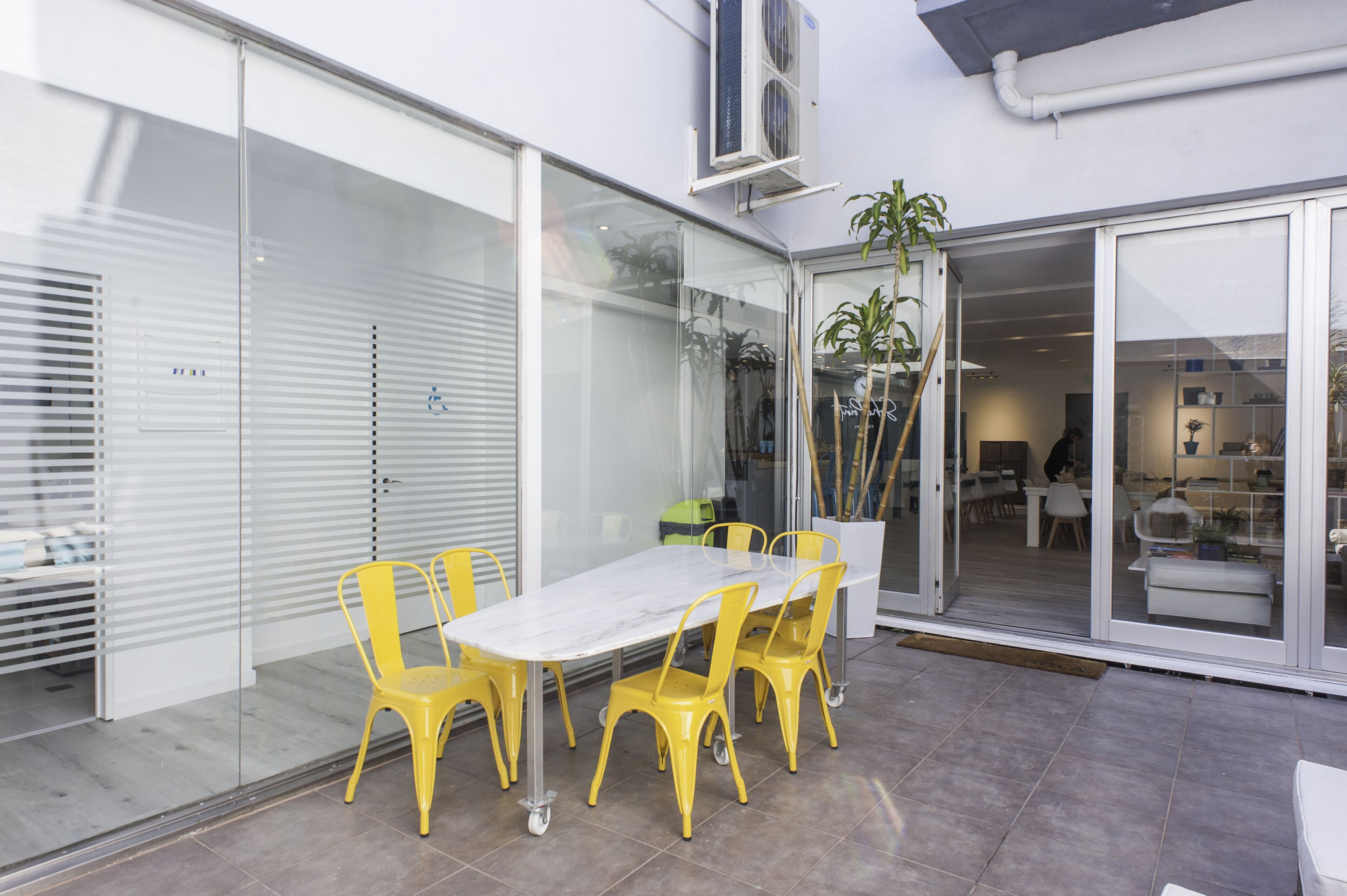 Un patio con una mesa blanca, sillas amarillas, una puerta corrediza de vidrio y una planta en maceta.