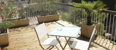 Terraza de madera con una mesa y dos sillas, rodeada de plantas en macetas y una barandilla de metal.