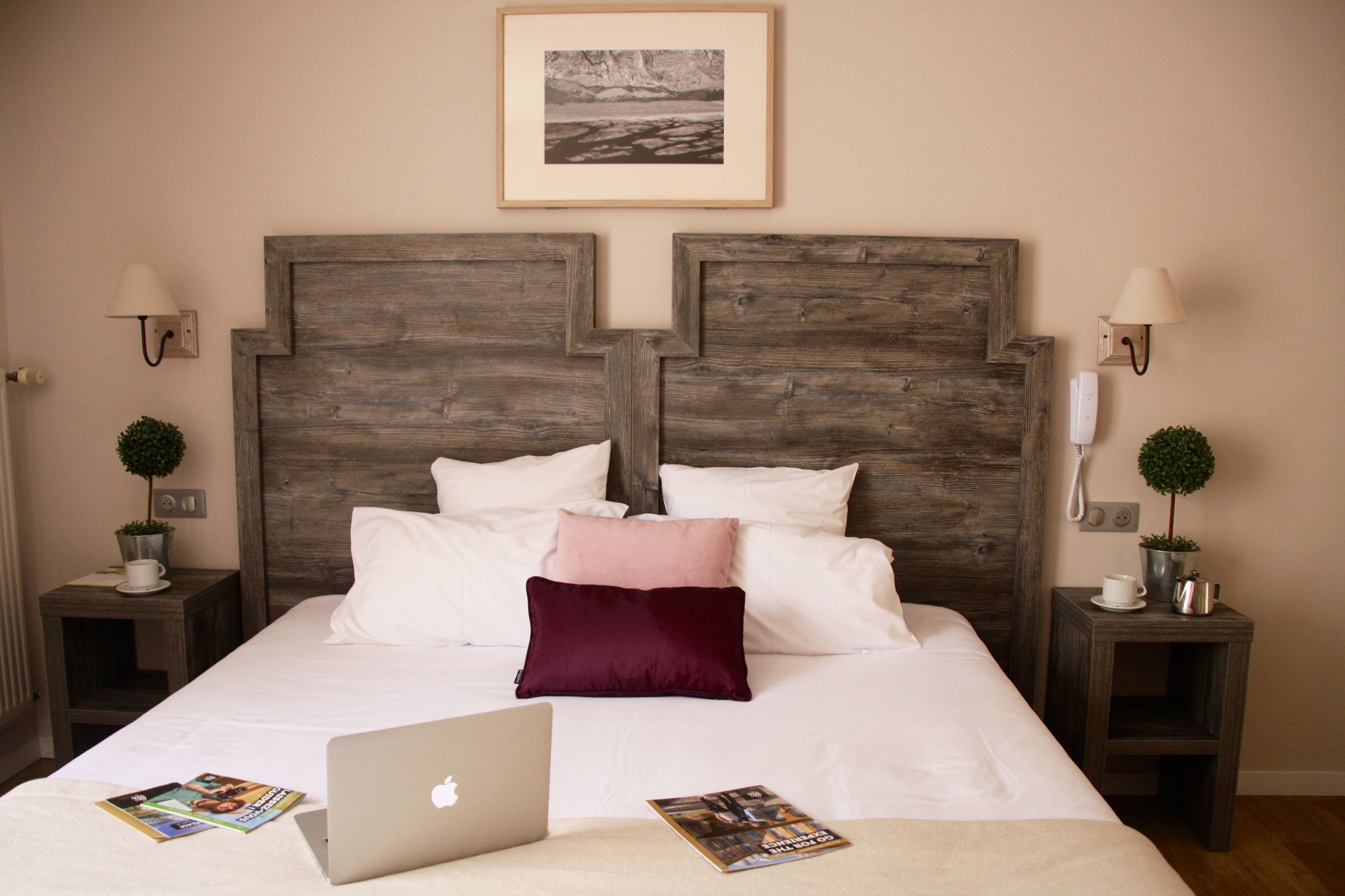 A neatly made bed with a laptop and magazines on it, wooden headboard, bedside tables with lamps and small potted plants, and a framed picture on the wall.