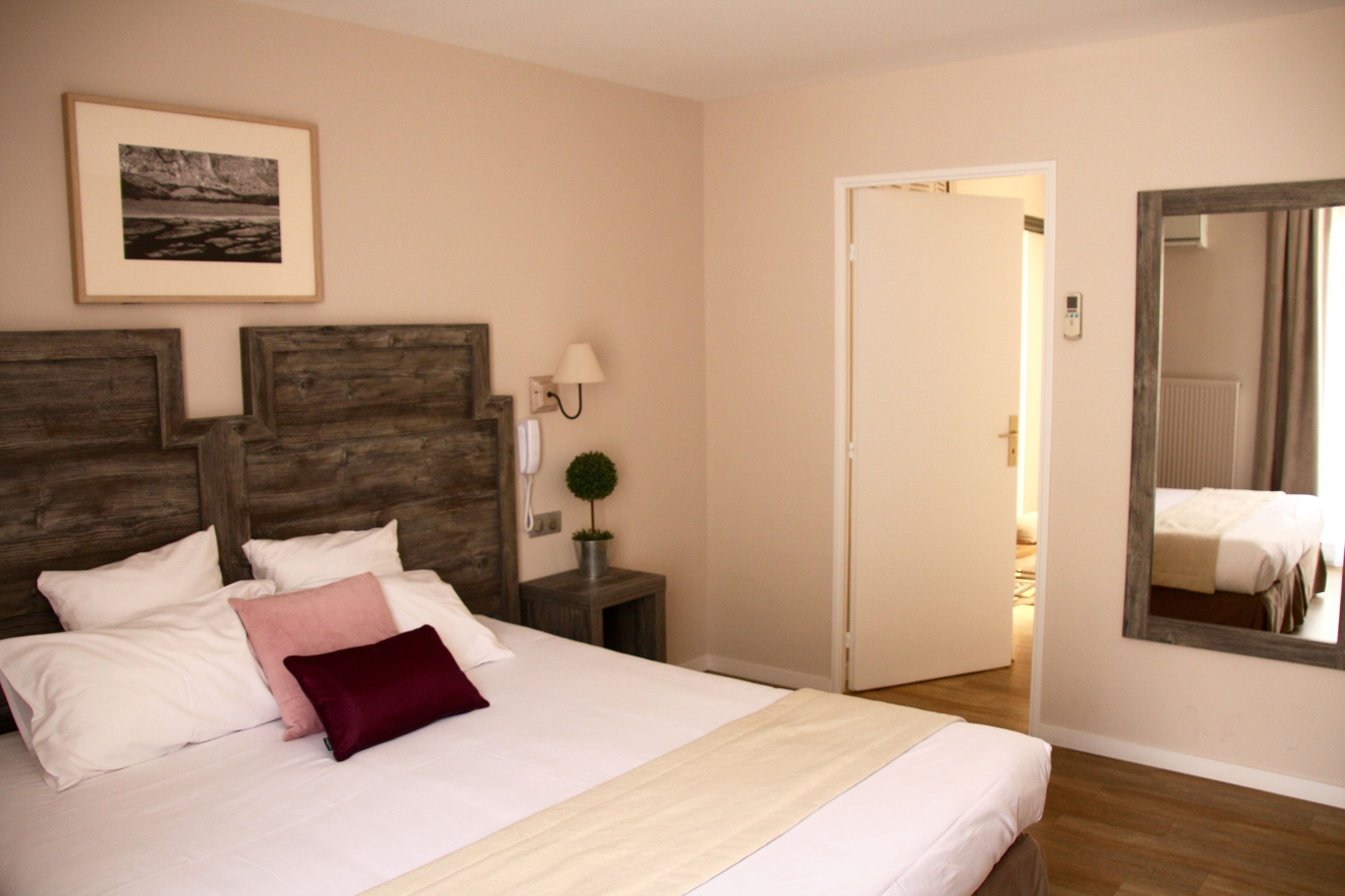 A bedroom with a large bed, a wooden headboard, a bedside table with a lamp, a framed picture on the wall, and a mirror on the wall.