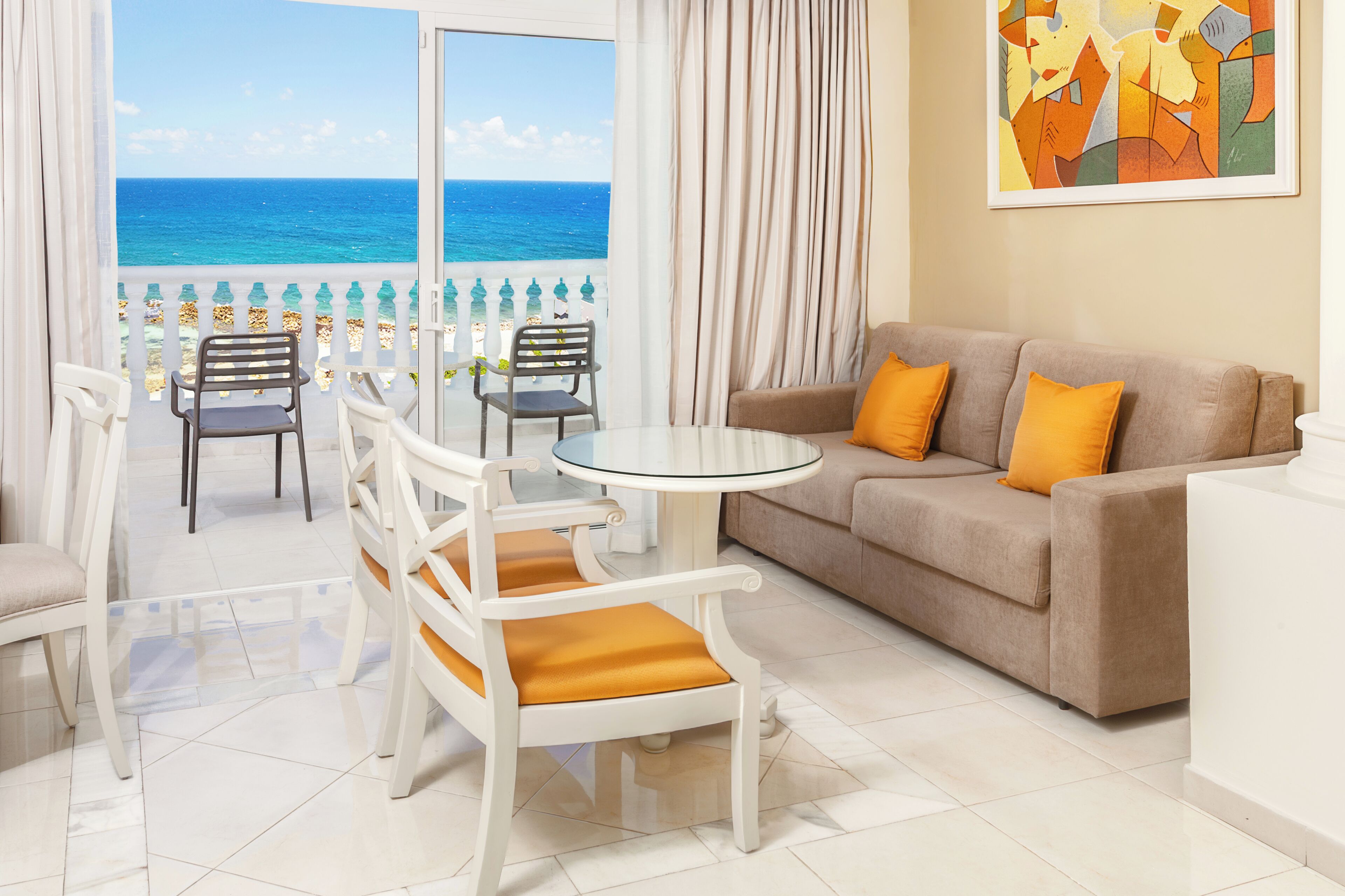 A room with a balcony, a sofa, two chairs, a round table, and a painting on the wall.