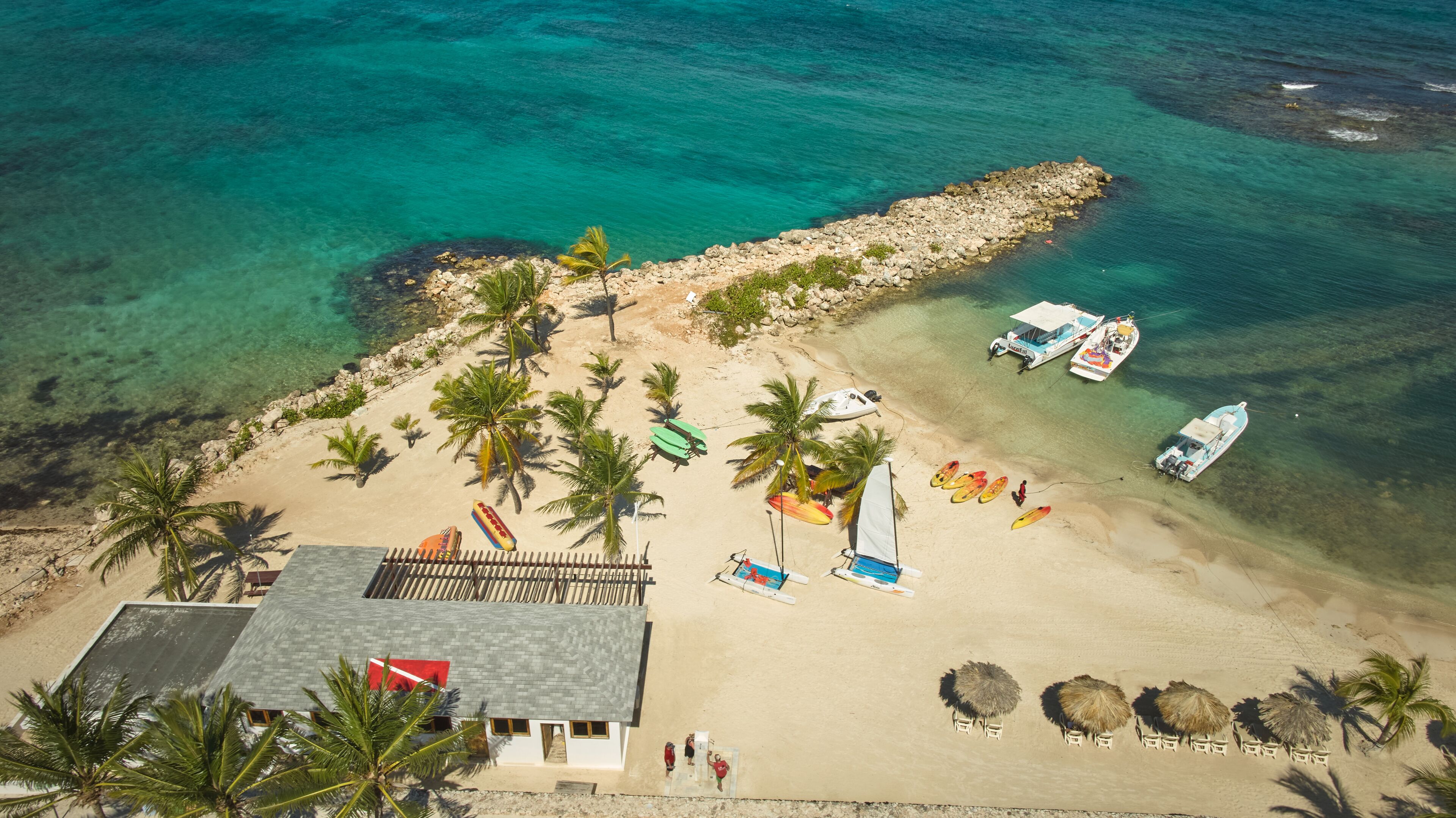 Luchtbeeld van een strand met helder turquoise water, een rotsachtige steenwal en verschillende boten afgemeerd dichtbij de kust. Er staan palmbomen, strandparasols en een gebouw met een schuin dak.