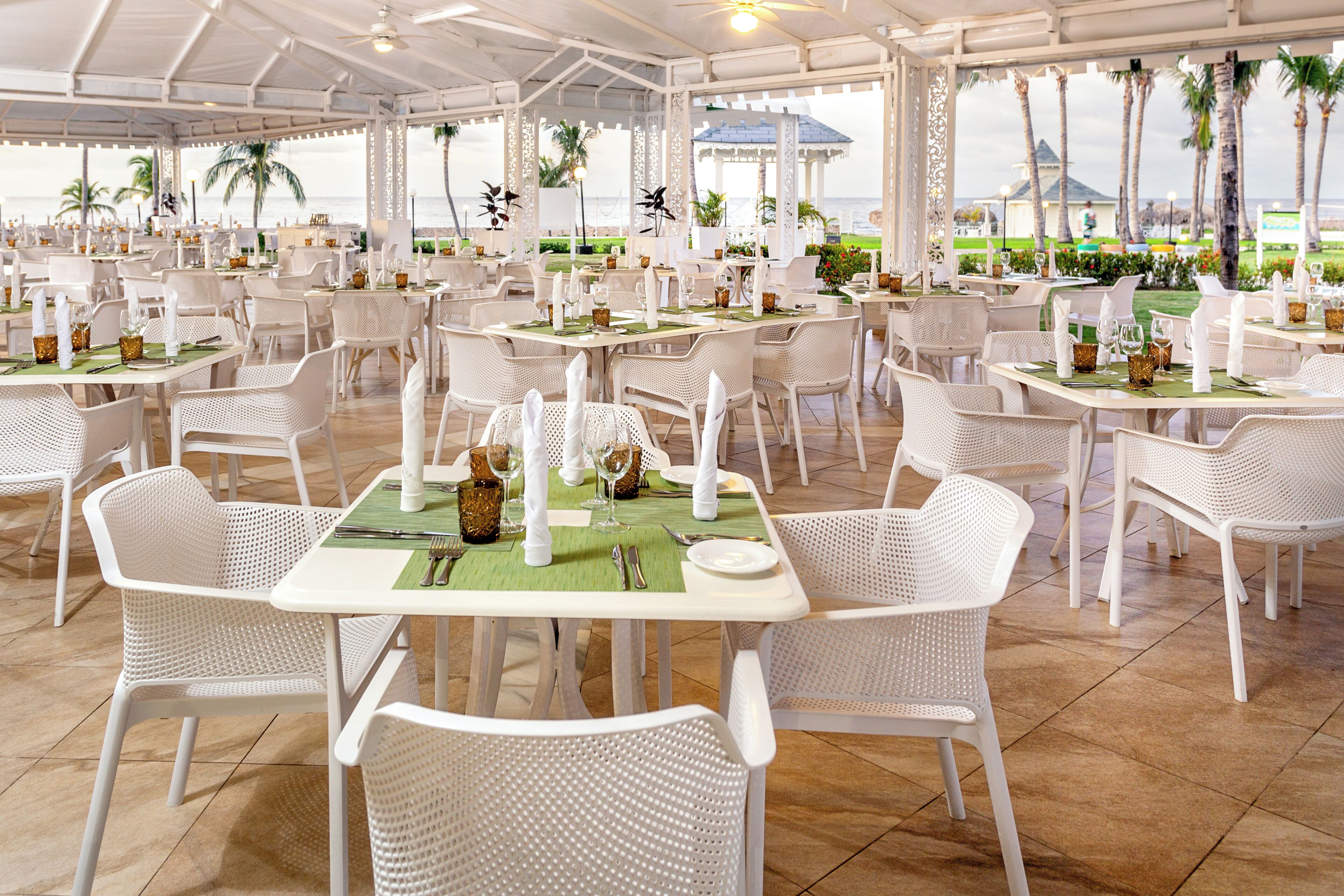 A spacious outdoor dining area with white wicker chairs and tables, arranged in a grid pattern, overlooking a scenic view of the ocean and palm trees.