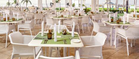 A spacious outdoor dining area with white wicker chairs and tables, arranged in a grid pattern, overlooking a scenic view of the ocean and palm trees.