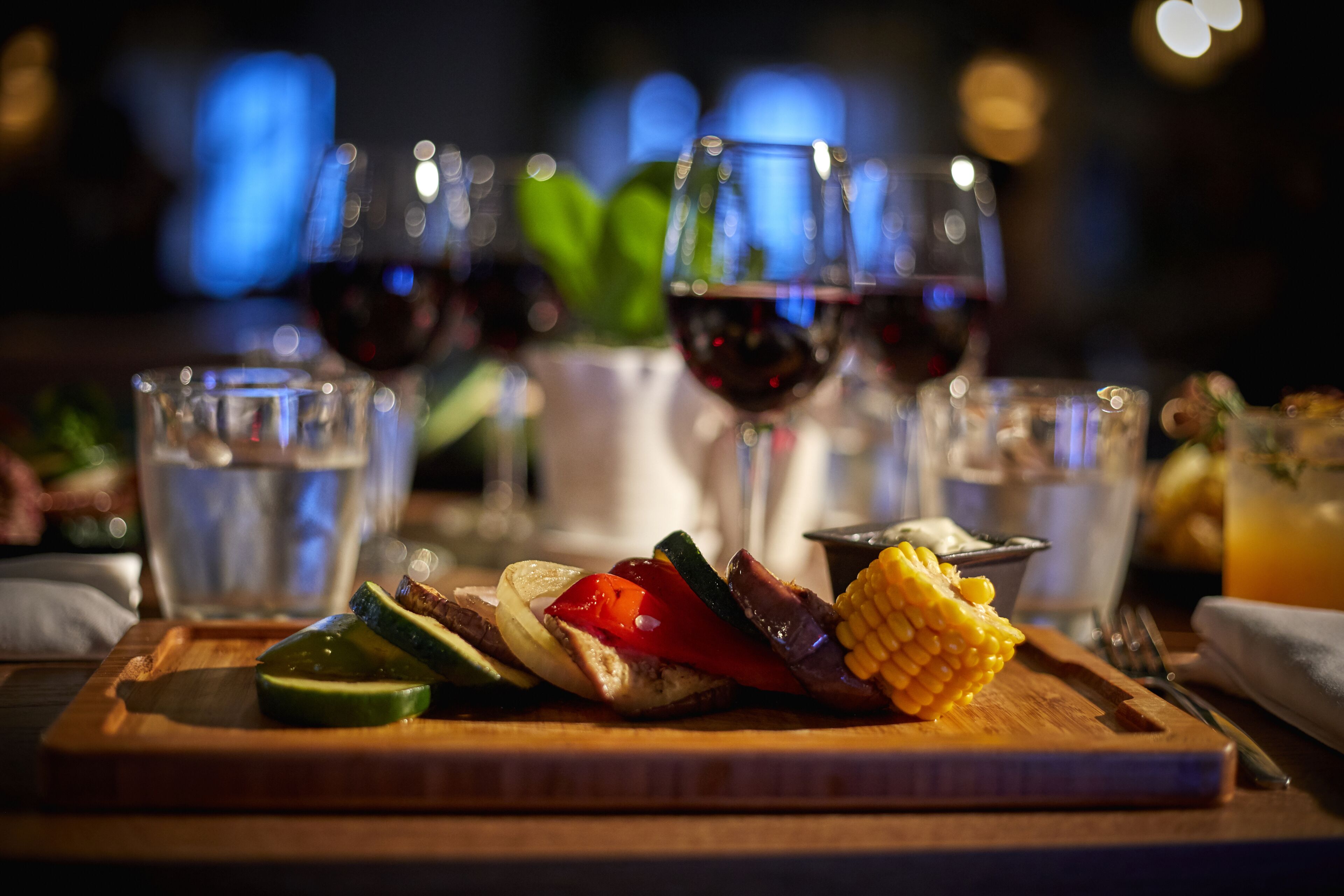 Une planche en bois garnie de légumes grillés et d’un épi de maïs, accompagnée de verres de vin rouge et d’un verre d’eau.