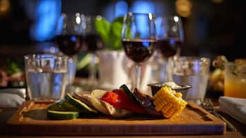 Une planche en bois garnie de légumes grillés et d’un épi de maïs, accompagnée de verres de vin rouge et d’un verre d’eau.
