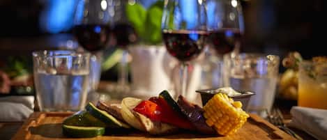 A wooden board with grilled vegetables and a corn cob, accompanied by glasses of red wine and a glass of water.