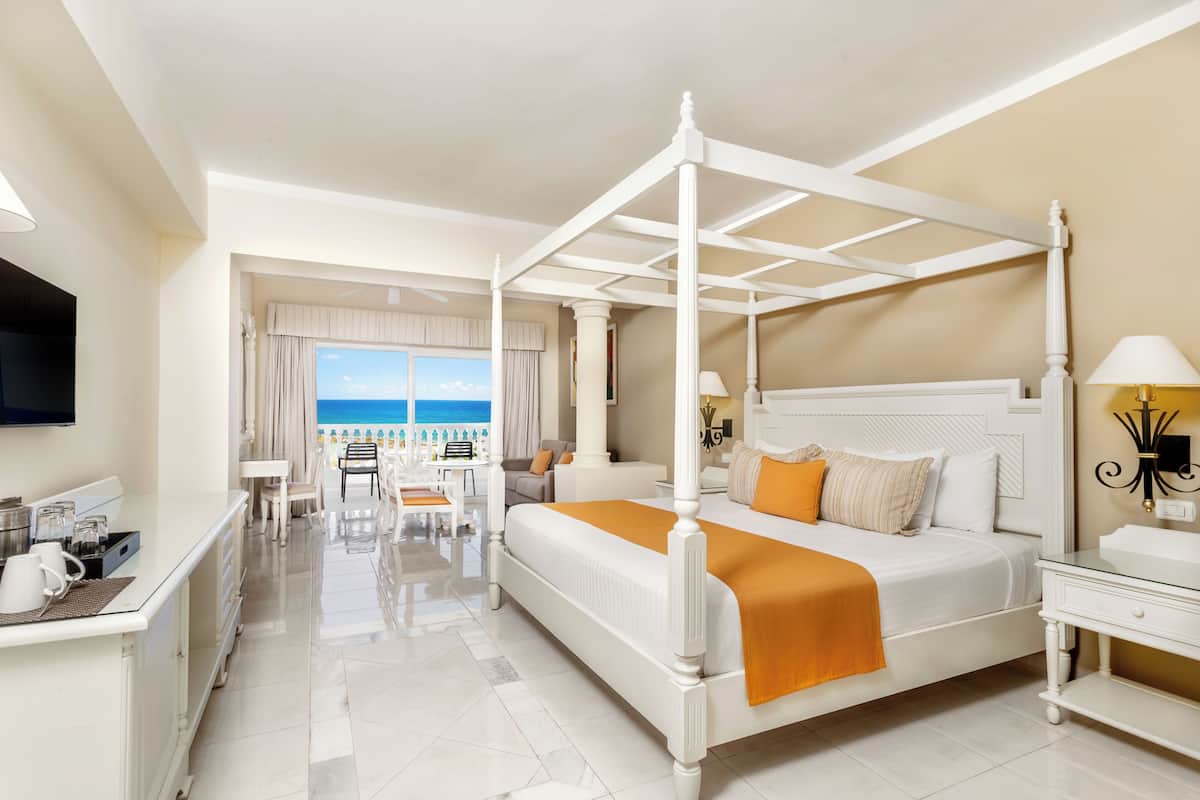 A four-poster bed with a white finish, a bedside table with a lamp, a flat-screen TV mounted on the wall, and a view of the ocean through a large window.