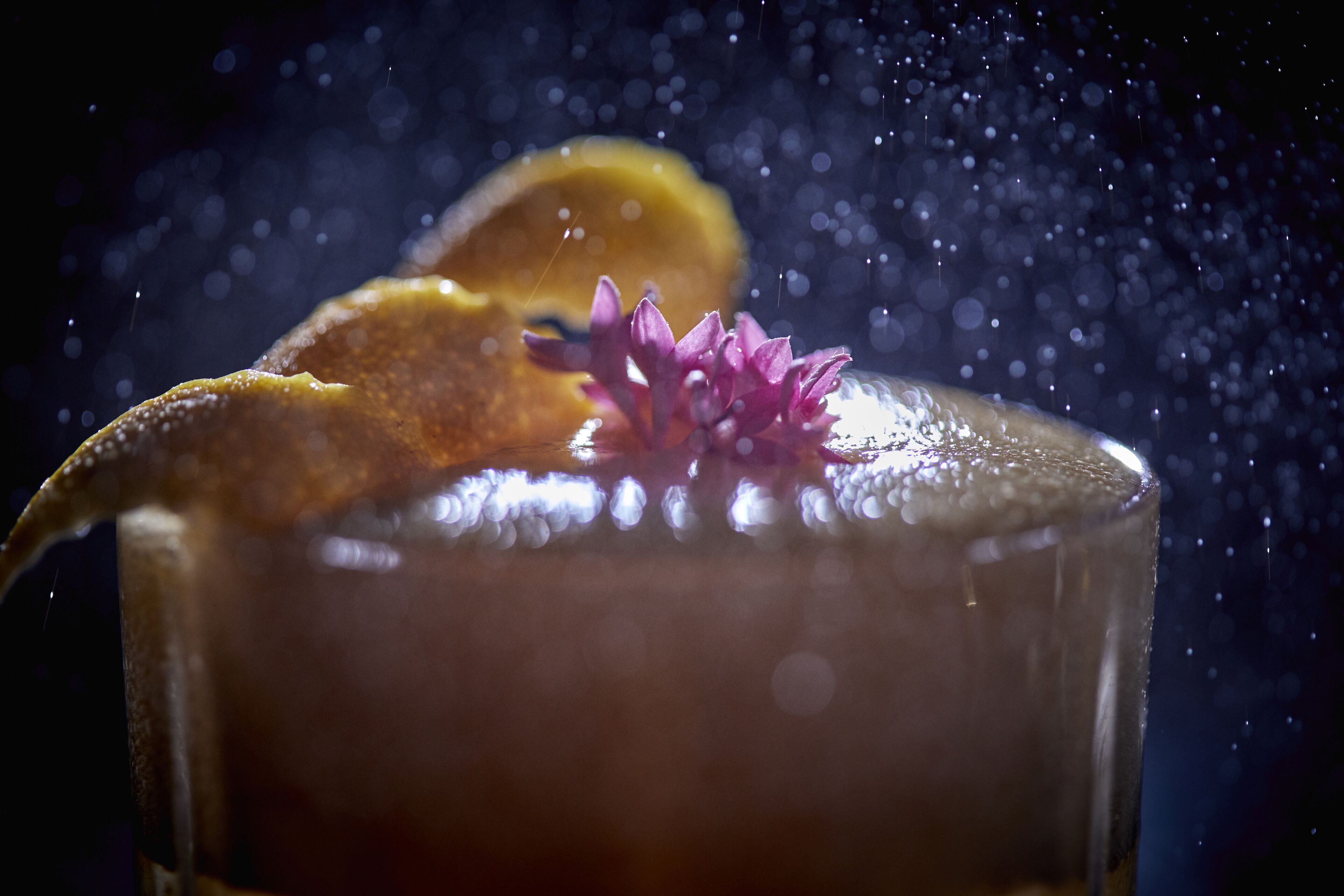 A close-up of a glass with a dark liquid, garnished with a slice of orange and a purple flower, with water droplets falling into it.