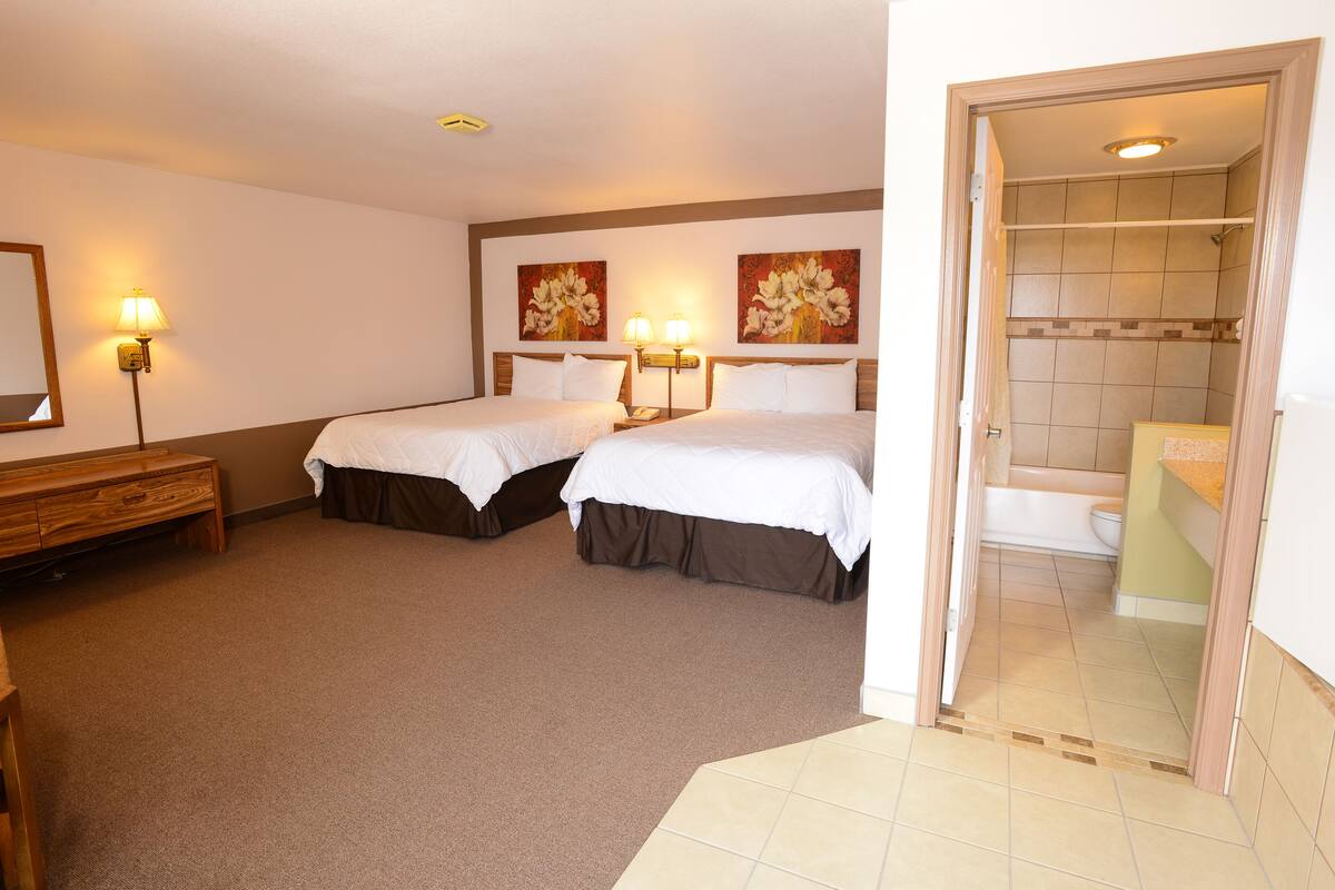 A hotel room with two beds, a wooden bench, a bathroom with a bathtub, and two wall-mounted lamps.
