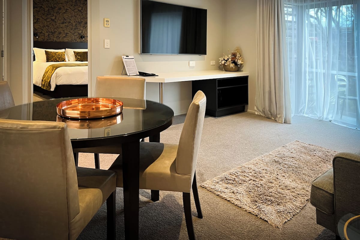 A modern hotel room with a bed, a desk, a TV, and a dining area with chairs and a table.