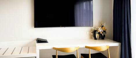 A modern room with a flat-screen TV mounted on the wall, a white desk with two wooden chairs, and a small potted plant on the desk.