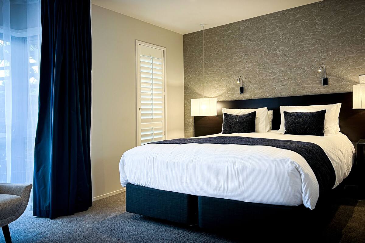A modern hotel room with a large bed, dark curtains, a wall-mounted lamp, and a patterned wall.