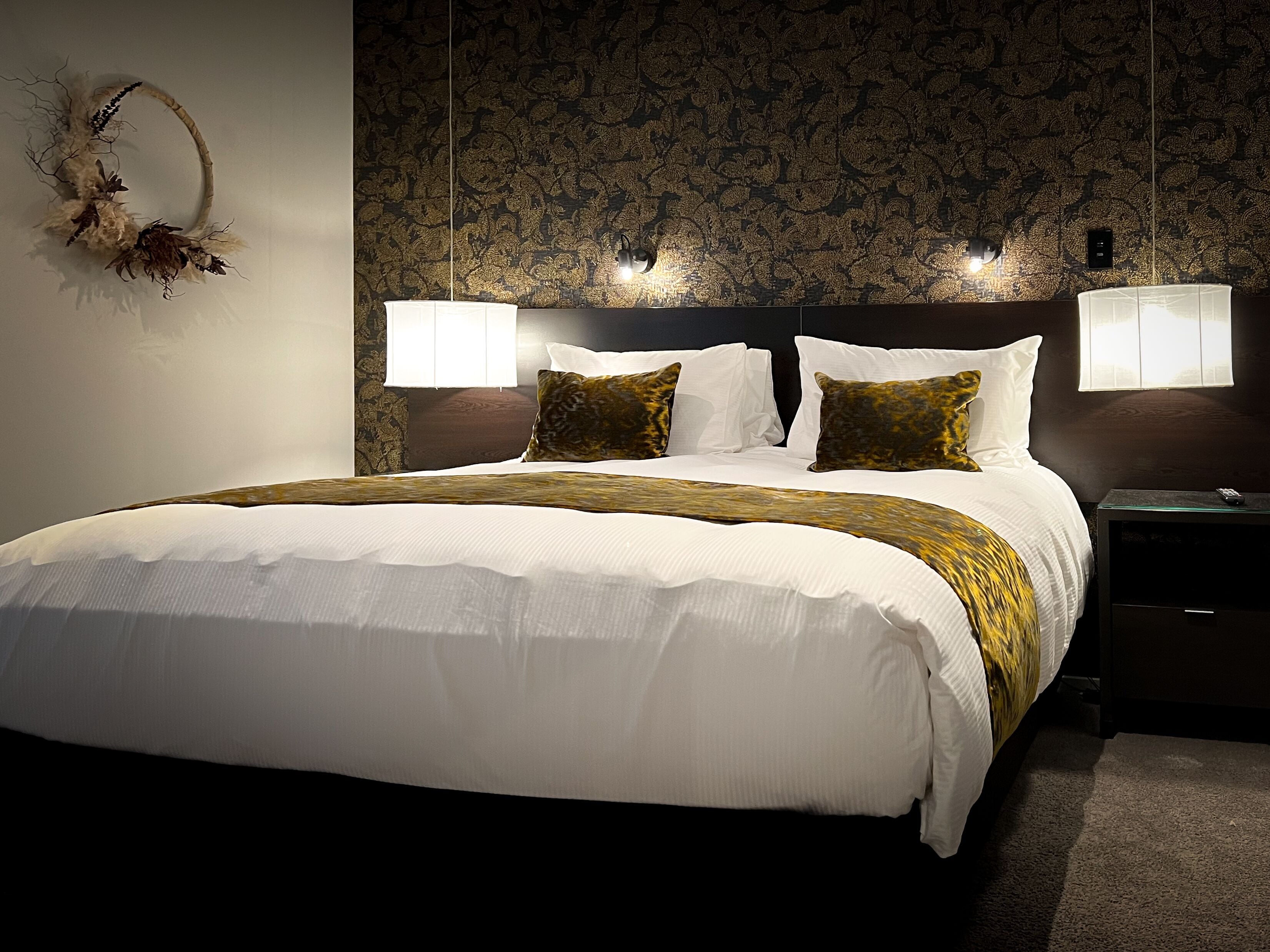 Una cama bien hecha con sábanas blancas y un cojín decorativo, un papel tapiz oscuro con diseño, y dos lámparas fijadas a la pared.