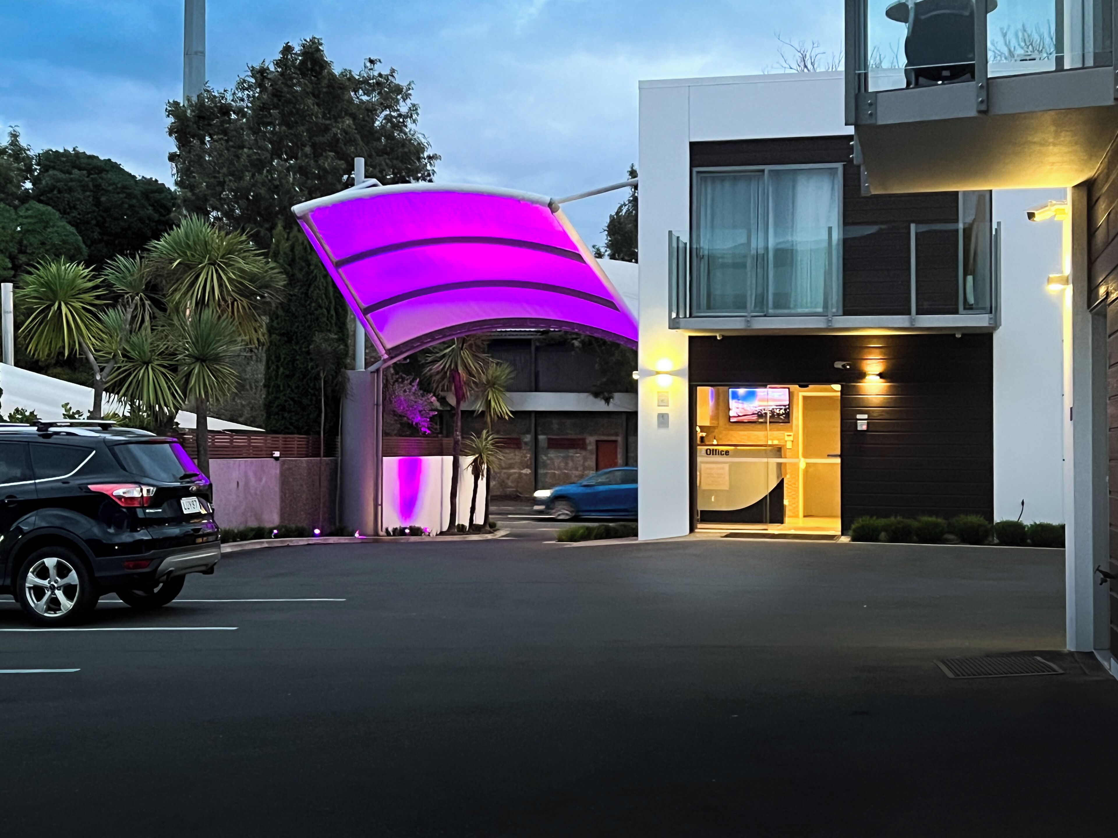 Un edificio moderno con un llamativo toldo morado, una recepción con televisor y un estacionamiento con un SUV negro.