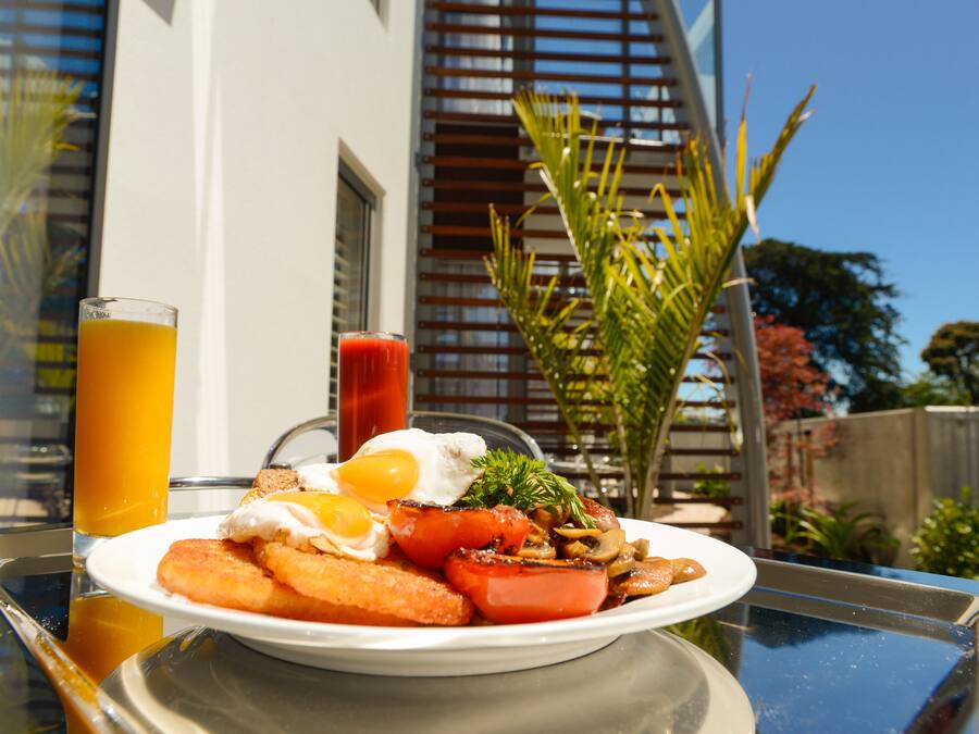 Un plato de desayuno con huevos, tomates y tostadas sobre una mesa frente a un edificio, con vistas a árboles y una cerca.