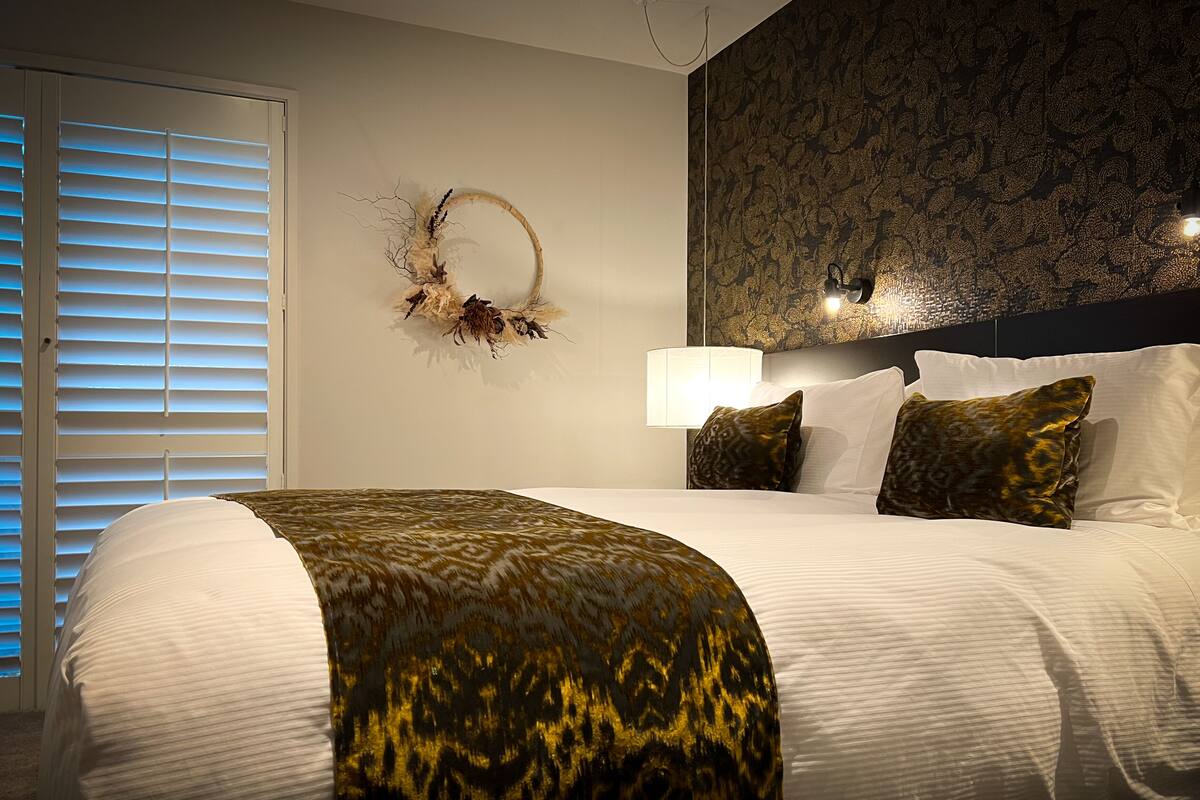 A neatly made bed with a patterned throw, a wall-mounted lamp, and a decorative wall piece.