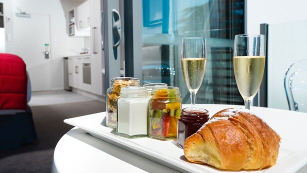 A breakfast tray with a croissant, fruit preserves, and two glasses of champagne on a white table in a modern hotel room with a view of a cityscape.