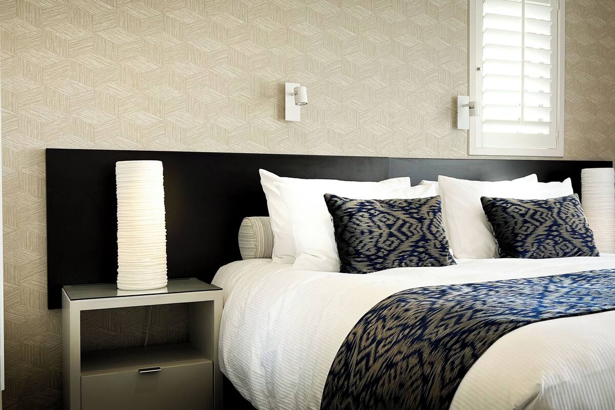 A neatly made bed with white and patterned pillows, a black headboard, and a bedside table with a lamp.