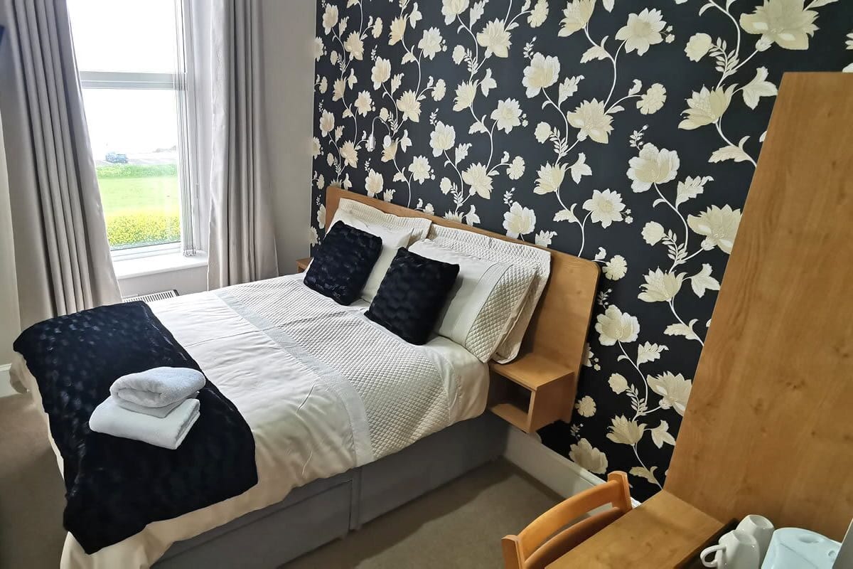 A neatly made bed with a white and black duvet, two pillows, and a folded towel on the side. A wooden nightstand is beside the bed. The room has a window with curtains, a wall with a floral pattern, and a small table with a cup and a book.