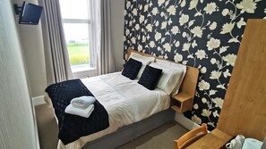 A neatly made bed with a white and black duvet, two pillows, and a folded towel on the side. A wooden nightstand is beside the bed. The room has a window with curtains, a wall with a floral pattern, and a small table with a cup and a book.