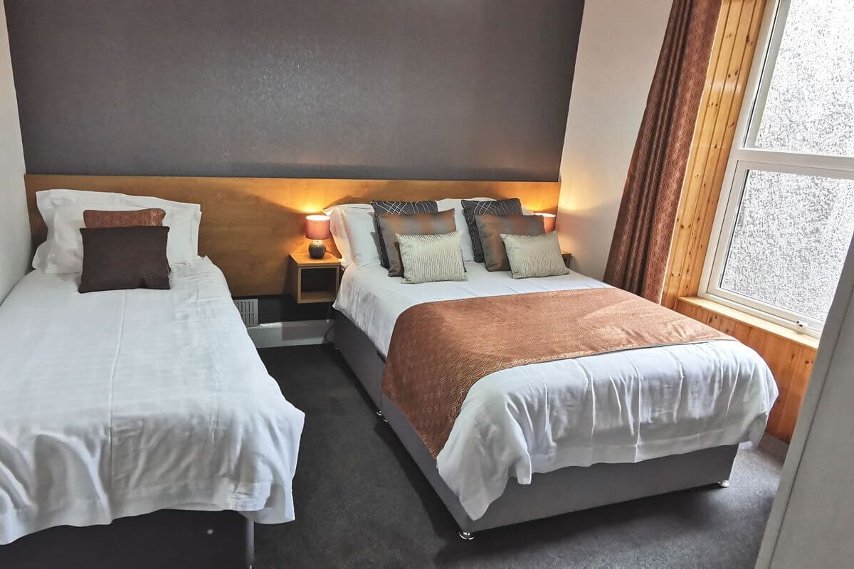 Two single beds with white linens, a wooden headboard, a nightstand with a lamp, and a window with curtains.