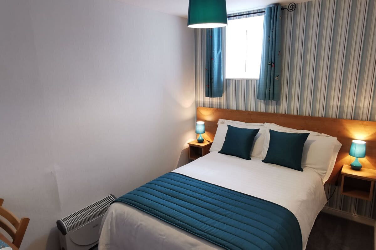 A neatly made bed with a blue bedspread and pillows, two bedside lamps, and a window with curtains.