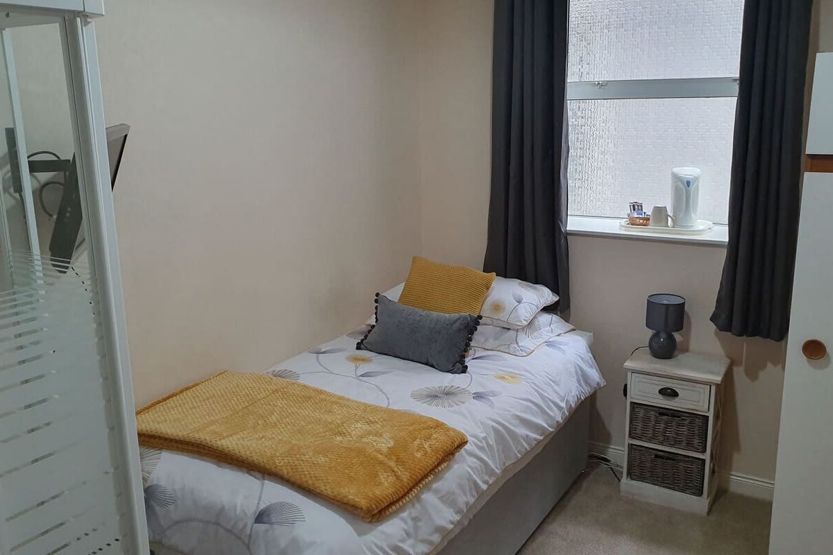 A small, neatly arranged bedroom with a bed, a nightstand, a window with curtains, and a wardrobe.
