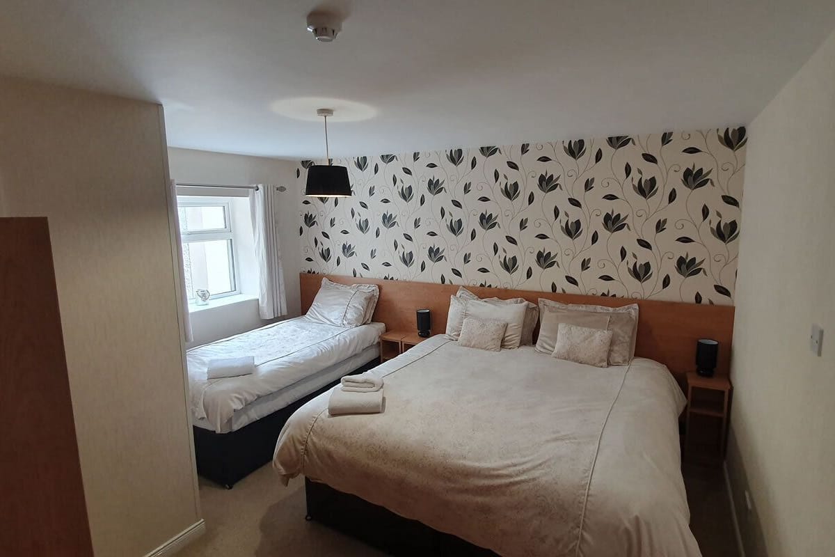 A bedroom with two single beds and one double bed, a wooden headboard, a patterned wallpaper, a ceiling light, and a window with curtains.