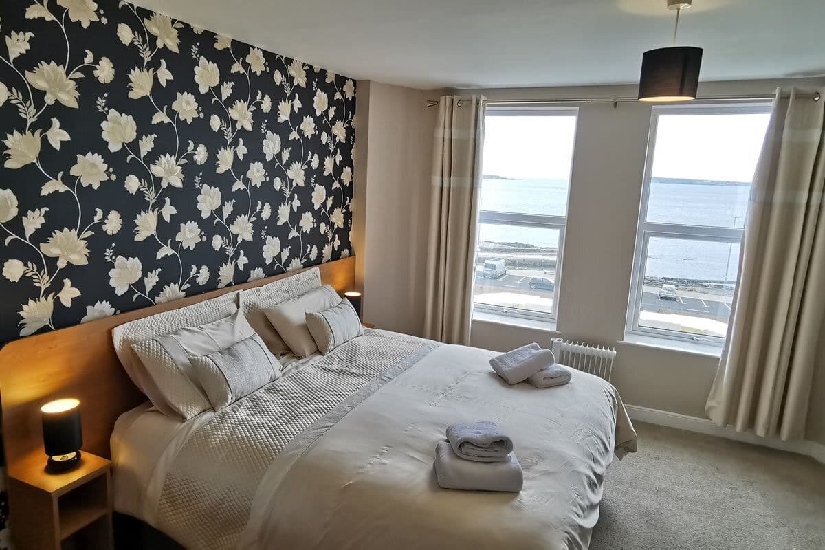 A hotel room with a large bed, a view of the sea, a window with curtains, and a bedside lamp.